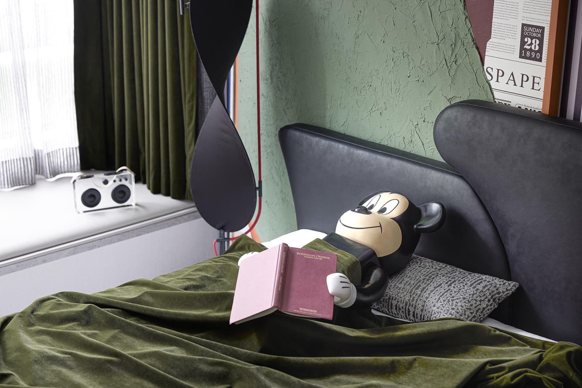 A modern bedroom with a black bed featuring a geometric pillow, green curtains, a green blanket, and a white speaker on the w