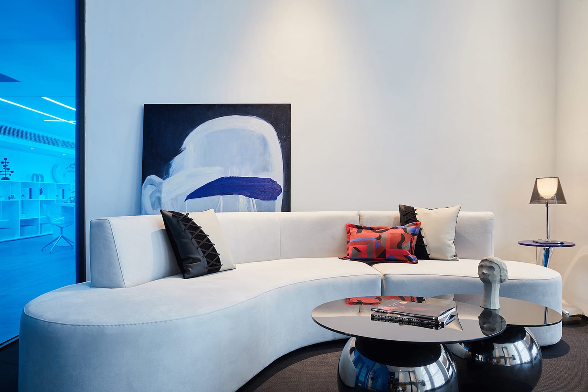 A modern minimalist living room featuring a white curved couch with colorful pillows, a round glass coffee table with books a