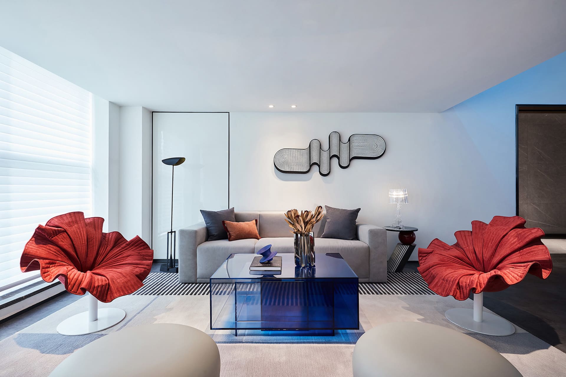 A modern living room features a sleek sofa with decorative pillows, minimalist red fabric chairs, a transparent blue coffee t