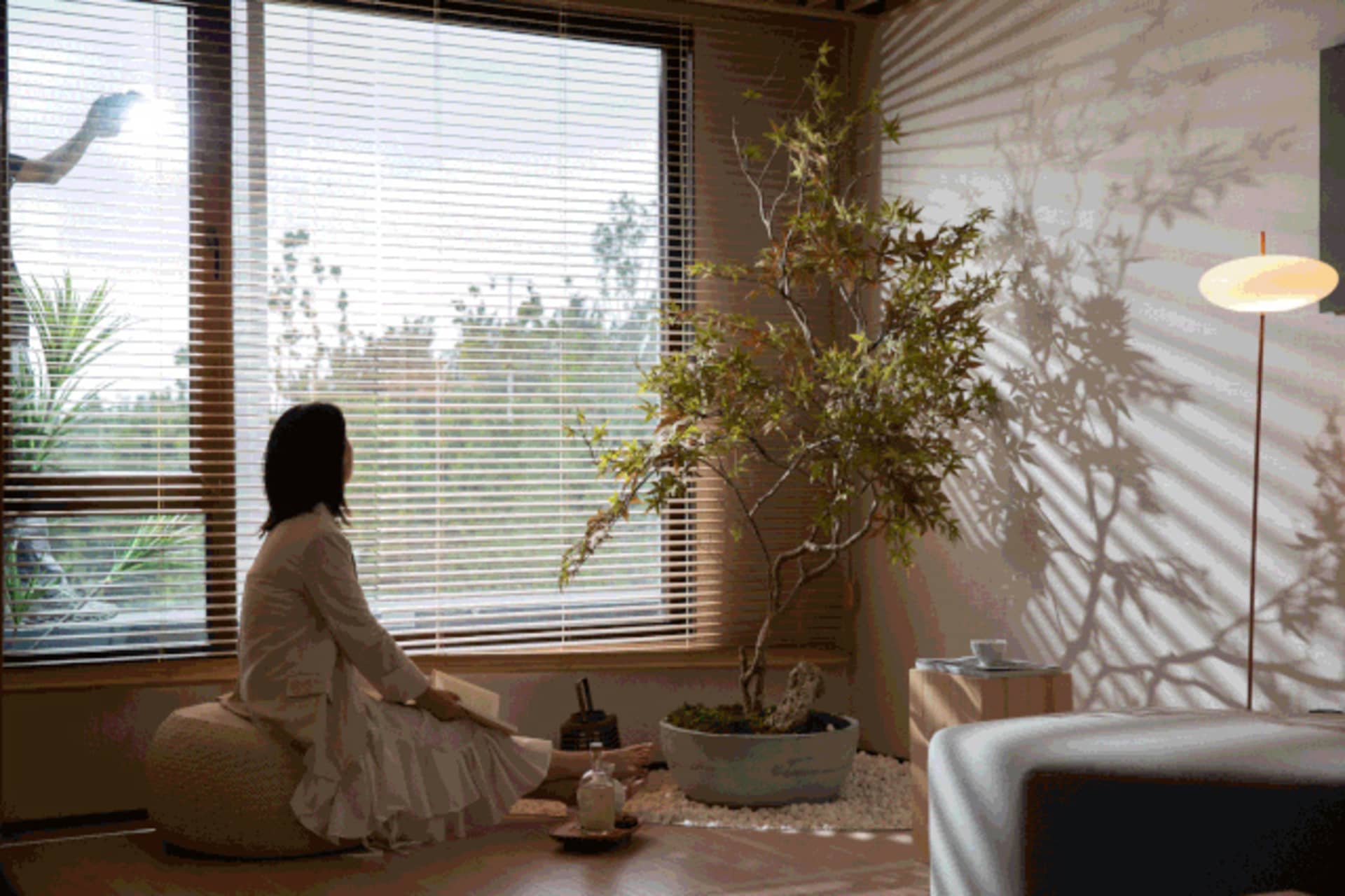 A woman sits on a floor cushion in a modern indoor living space with plants and a floor lamp.