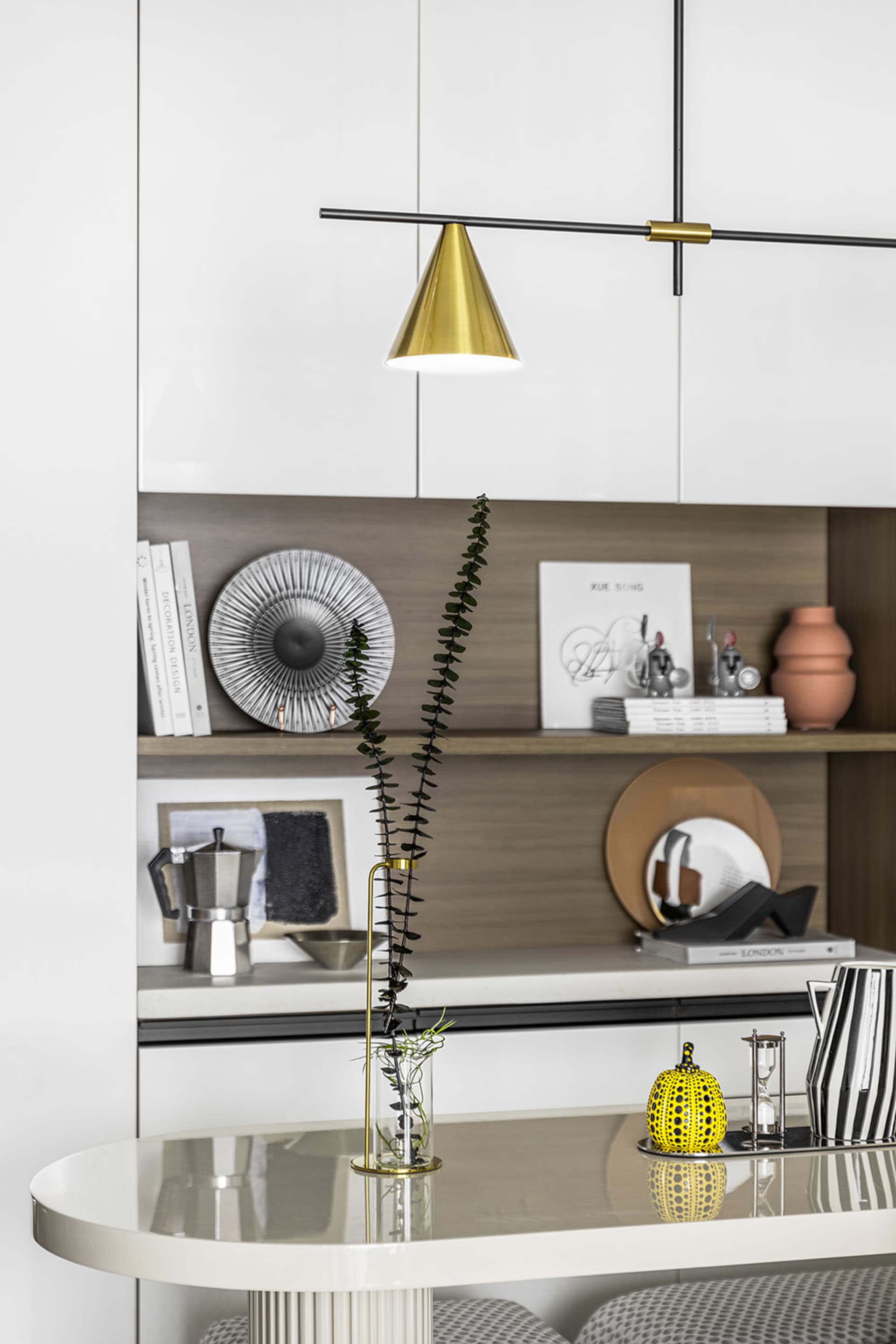 Modern kitchen with sleek white cabinets, metallic lighting fixture, and decorative vase on white table.