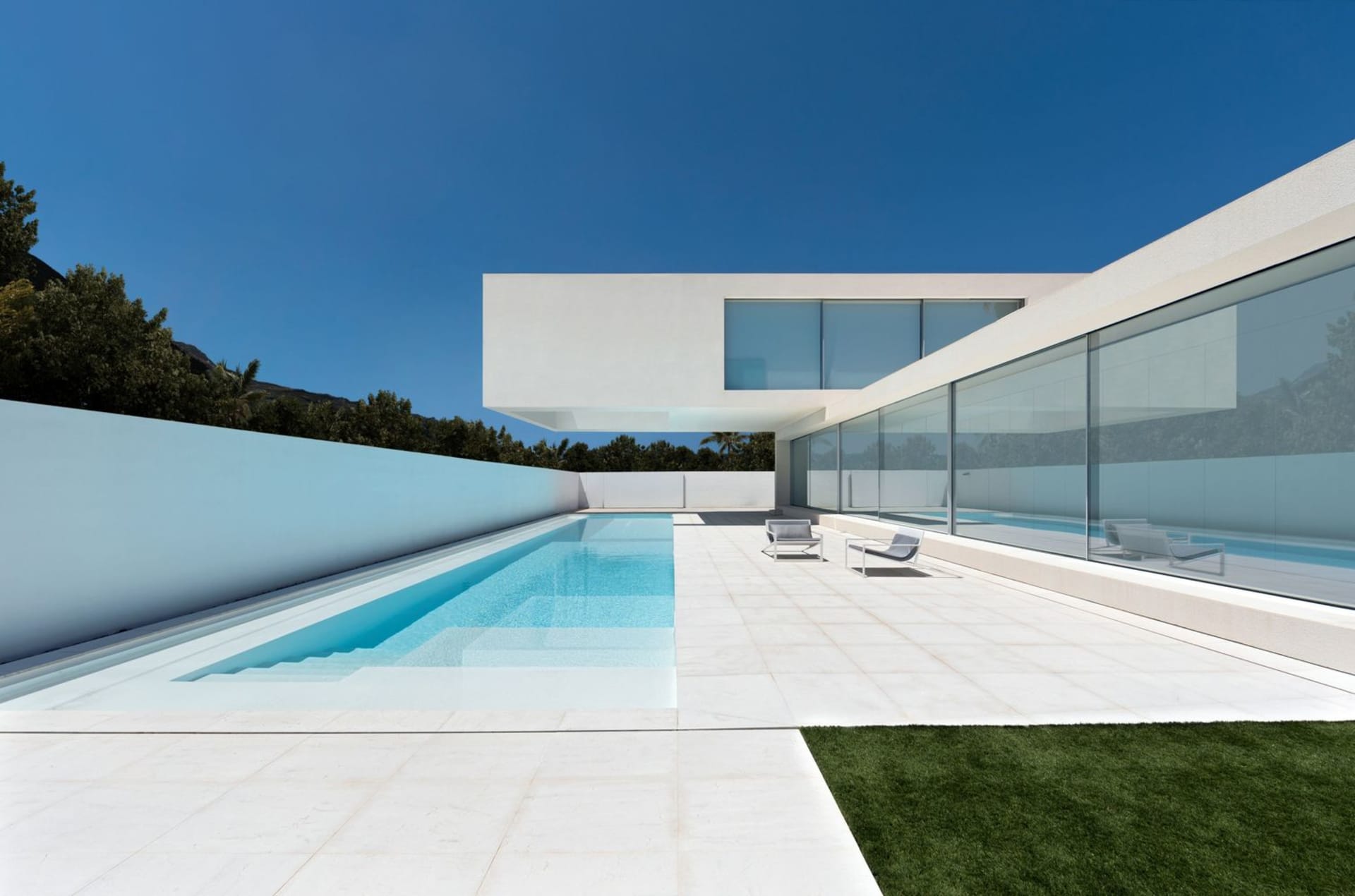 Modern minimalist residential exterior with concrete walls and large glass windows, featuring a sleek white patio area and a