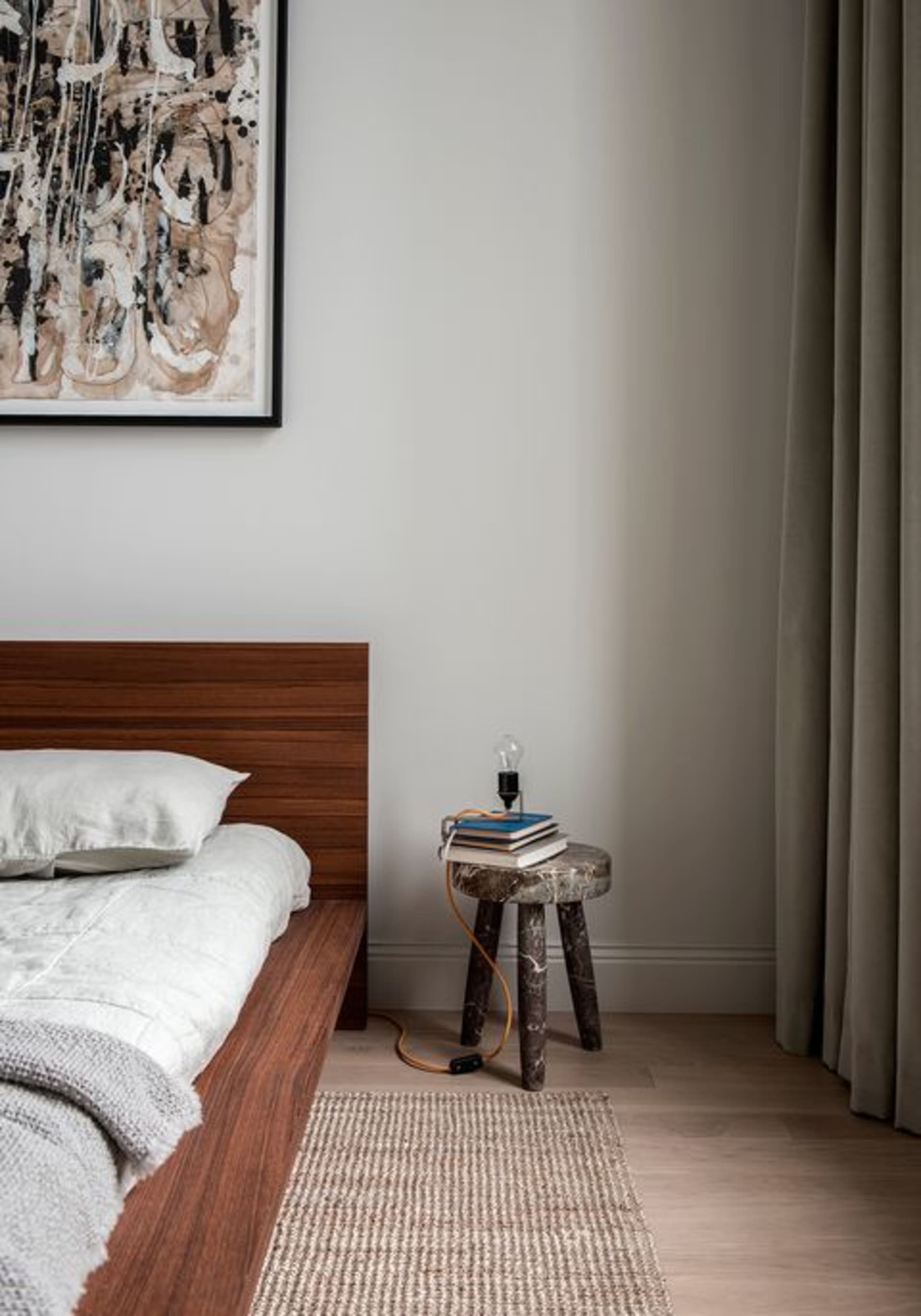 A minimalist bedroom featuring wooden furniture and a hanging light bulb on a stool, with a framed abstract painting above th