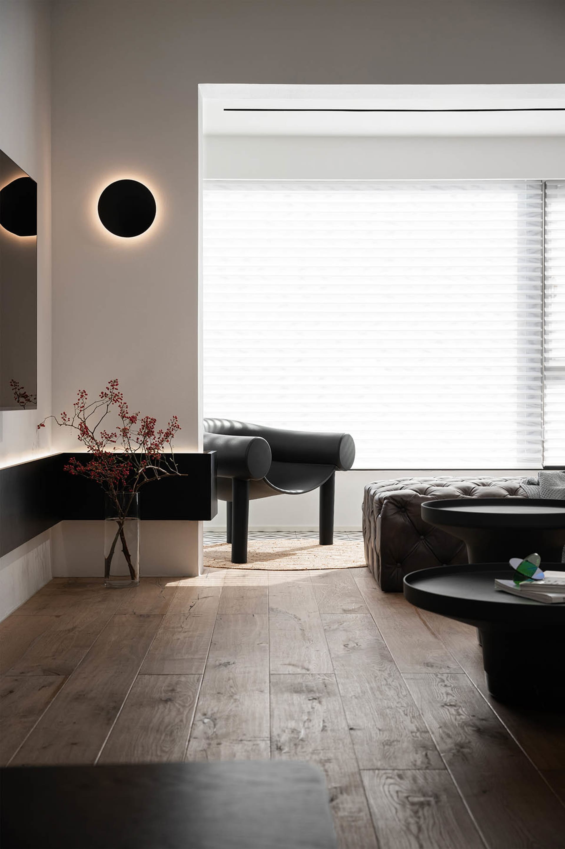 A modern minimalist living room featuring wooden floors, black furniture, and a large window with blinds.