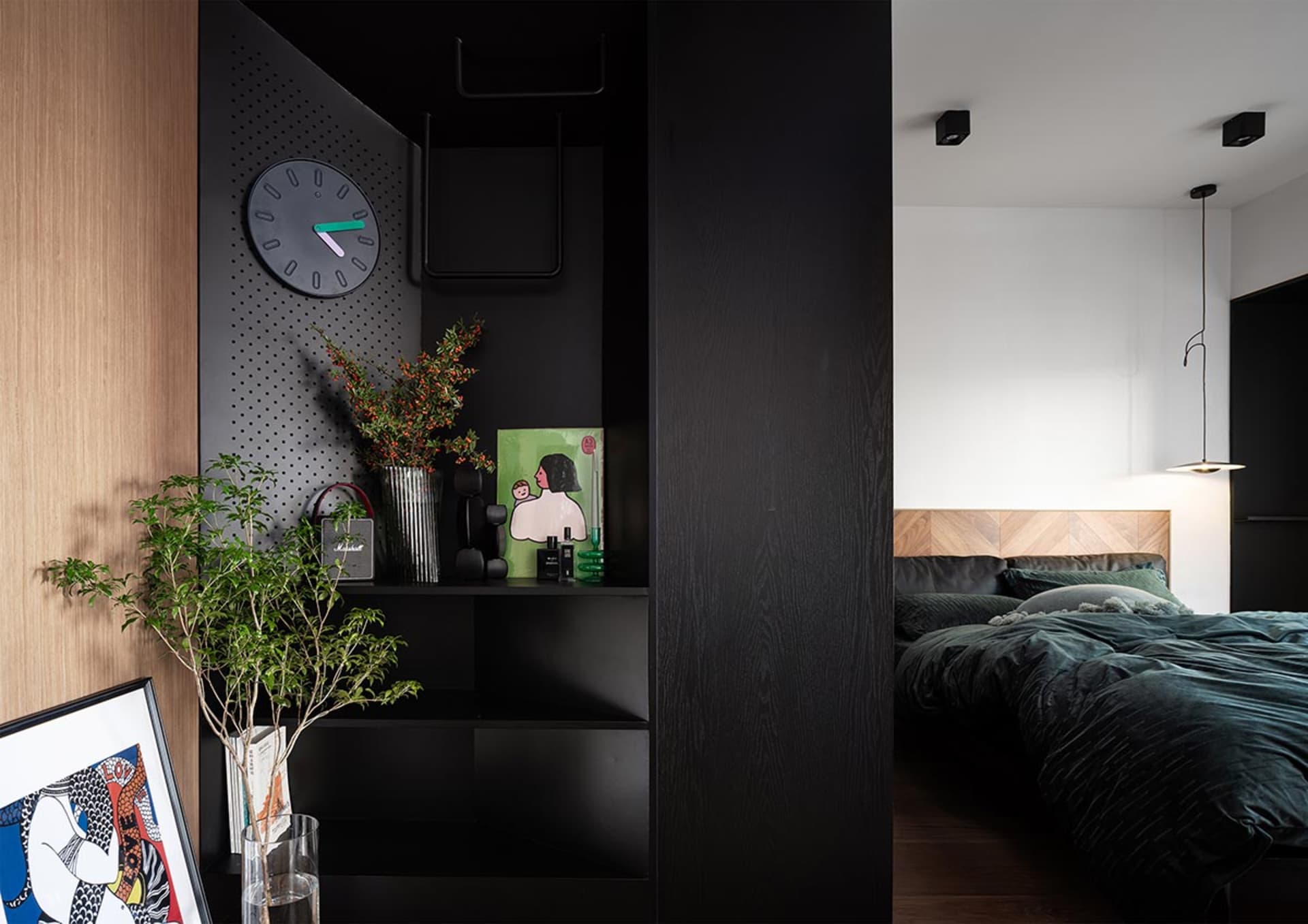 A modern bedroom featuring a dark-colored shelf with a clock, plants, and decor, leading to a bed with velvet bedding and a w