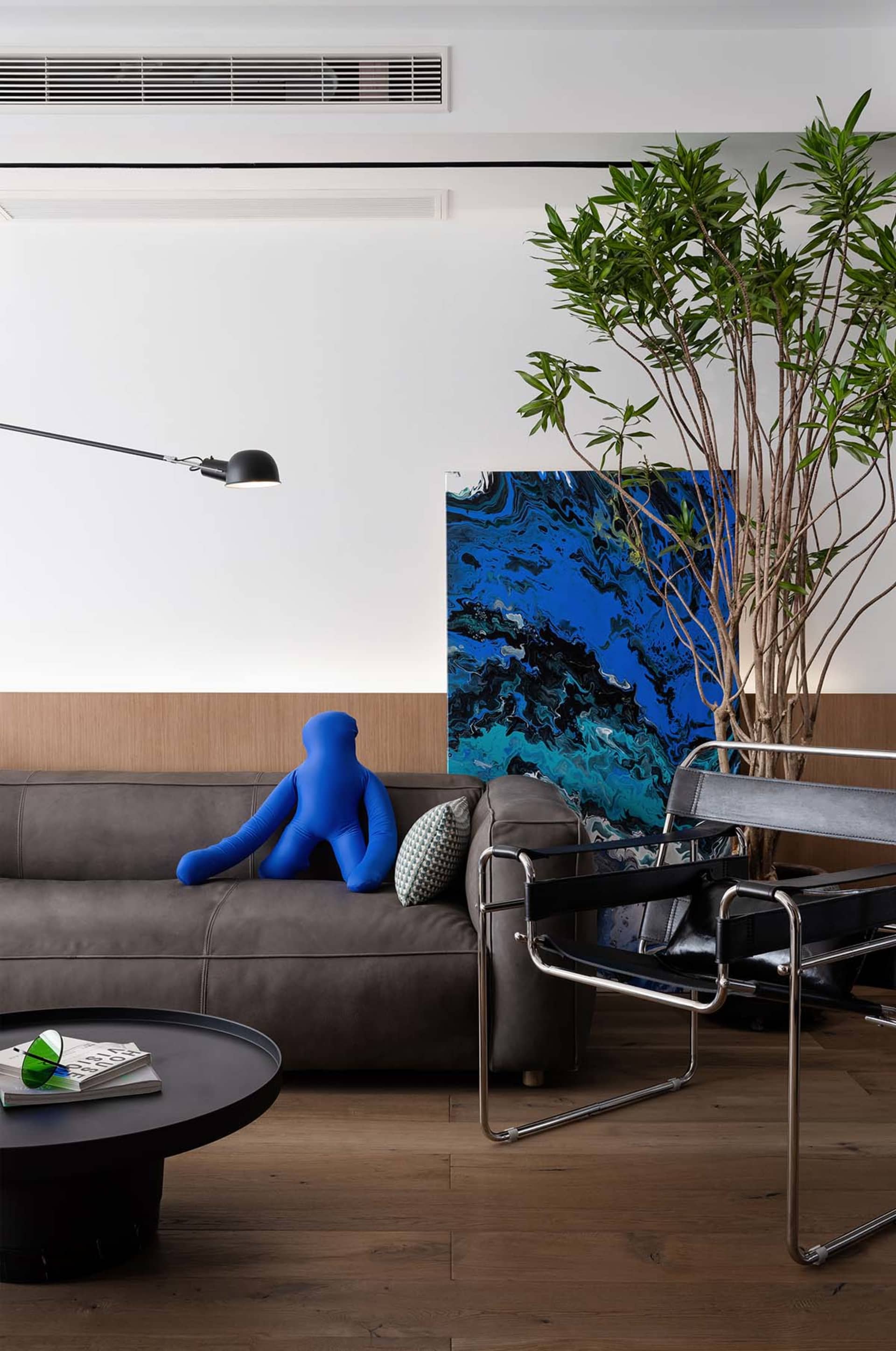Modern living room with a leather sofa, a blue figure, a black and blue painting, and a potted plant.