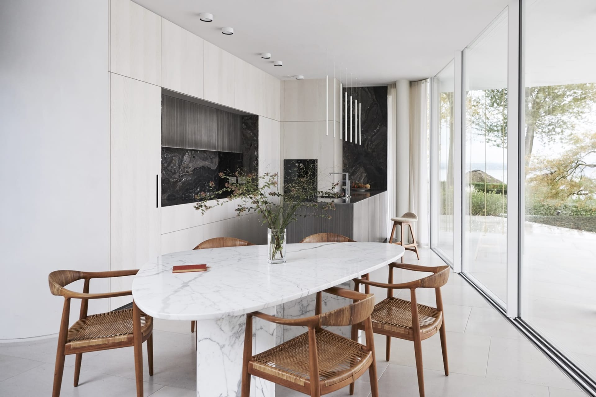 A modern residential kitchen featuring a white marble table and wooden chairs, with a view of the outdoors through large glas