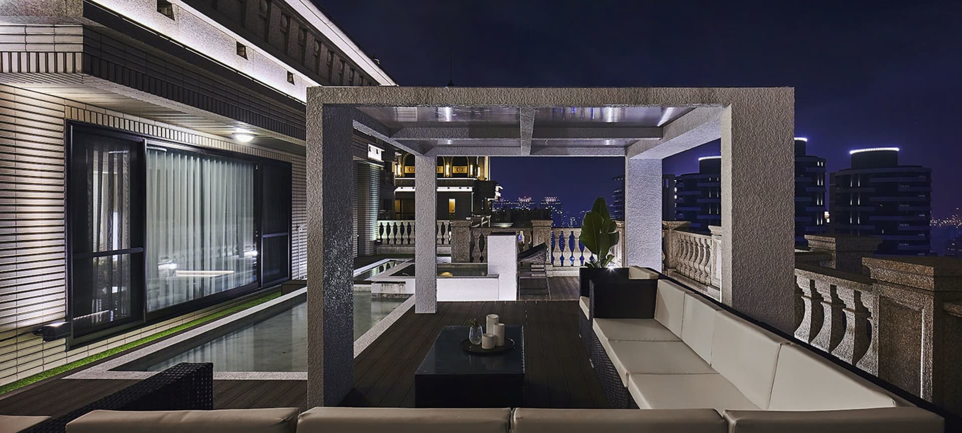 Modern rooftop patio with concrete and glass elements, featuring a lounge area and cityscape view at night.