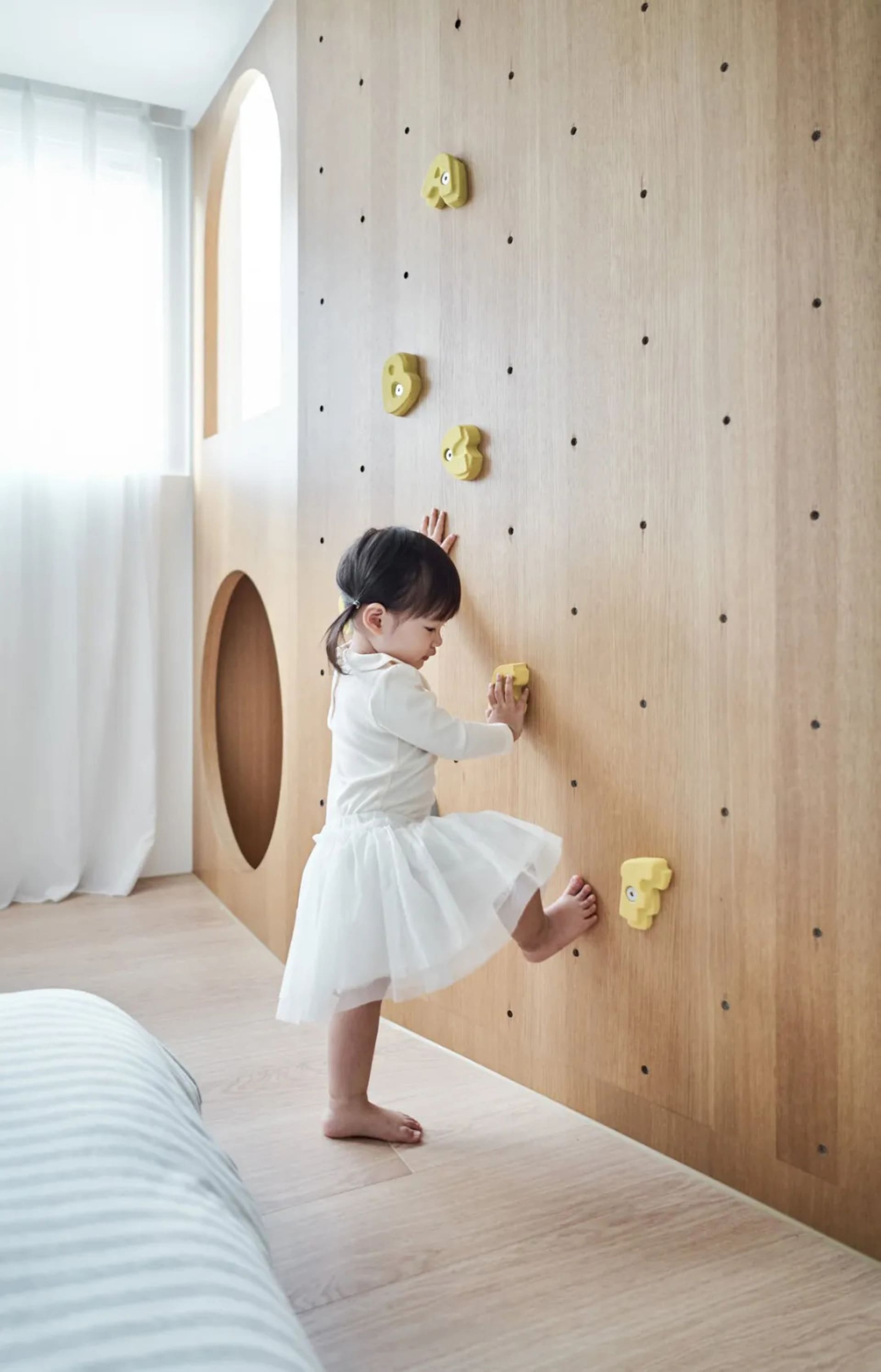 A toddler climbing on a wooden wall with yellow climbing holds in a modern children's playroom.