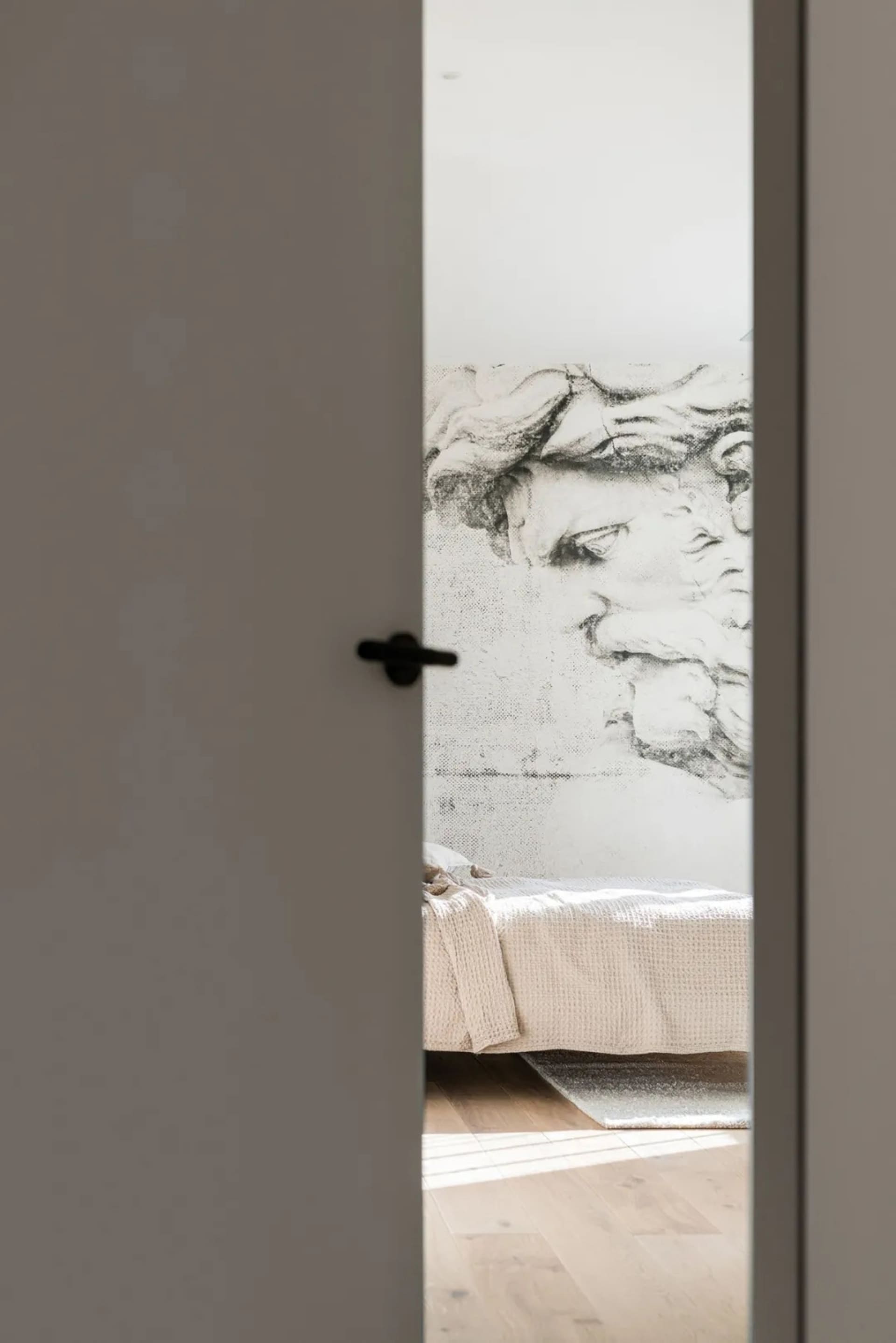 A minimalist bedroom featuring a concrete wall with a large, detailed wall mural of a classical statue's head, a bed with a t