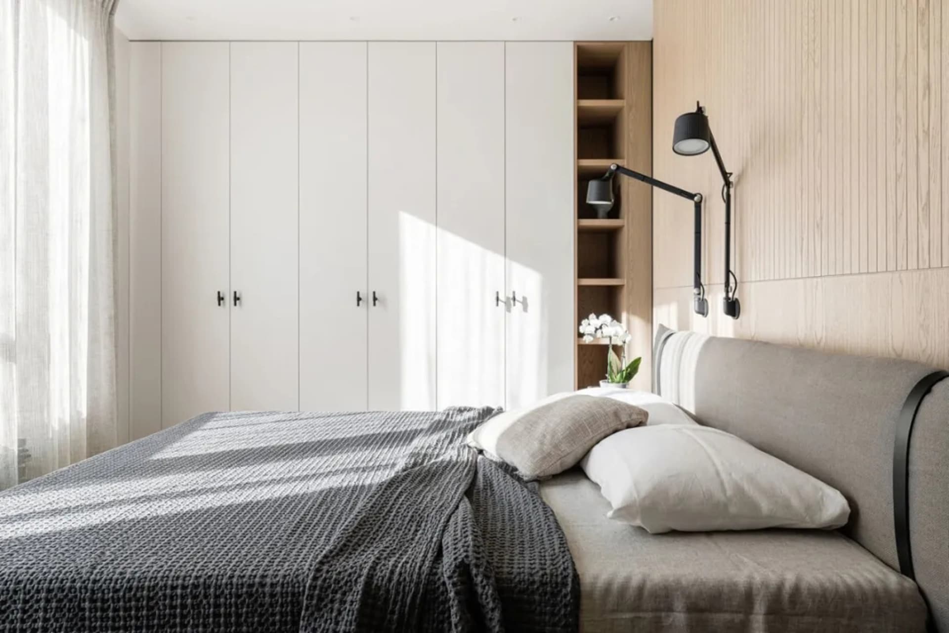 Modern minimalist bedroom featuring a grey upholstered bed against a wall of light wood panels and a large white wardrobe.