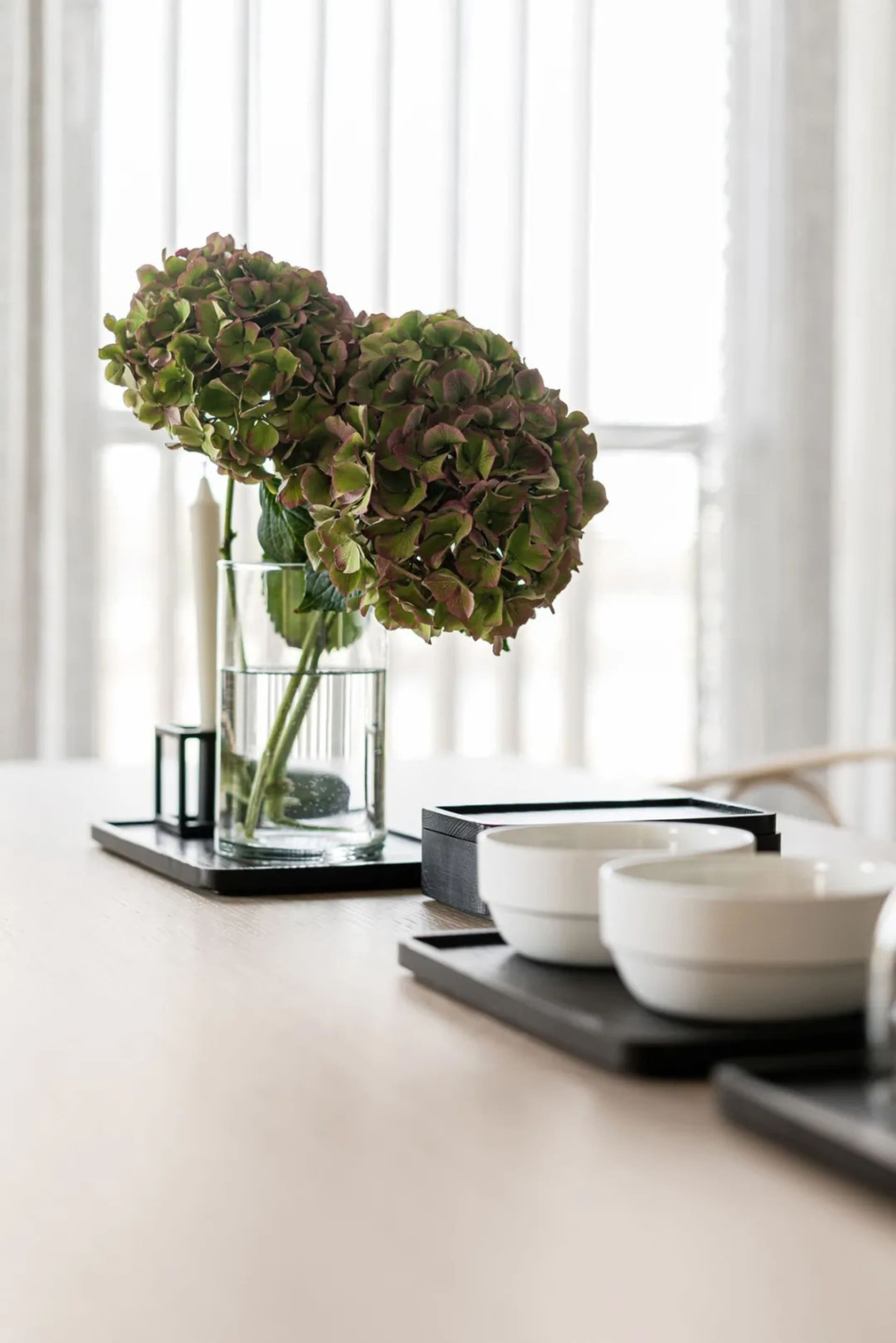 A minimalistic table setting with a clear glass vase containing hydrangea flowers and bowls on a tray.