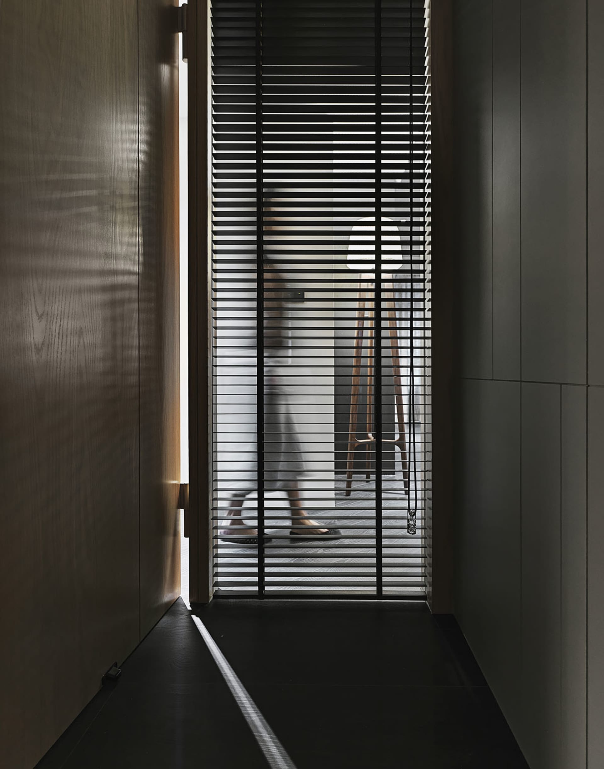A modern doorway with metal blinds partially open, showing a wooden floor and a person standing behind the blinds.