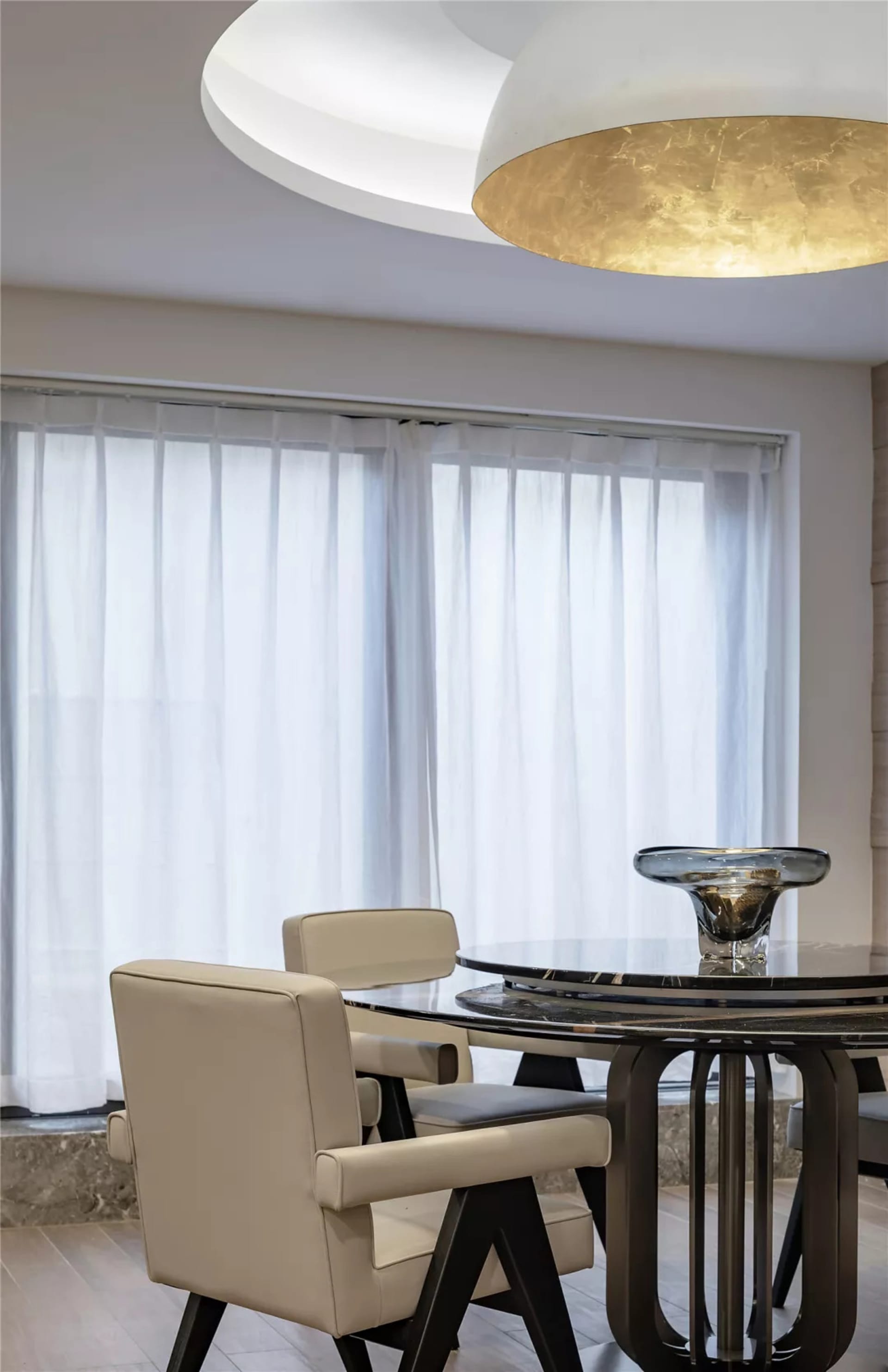 A modern dining room featuring a round glass-top table with black marble base, white leather upholstered chairs, and a large
