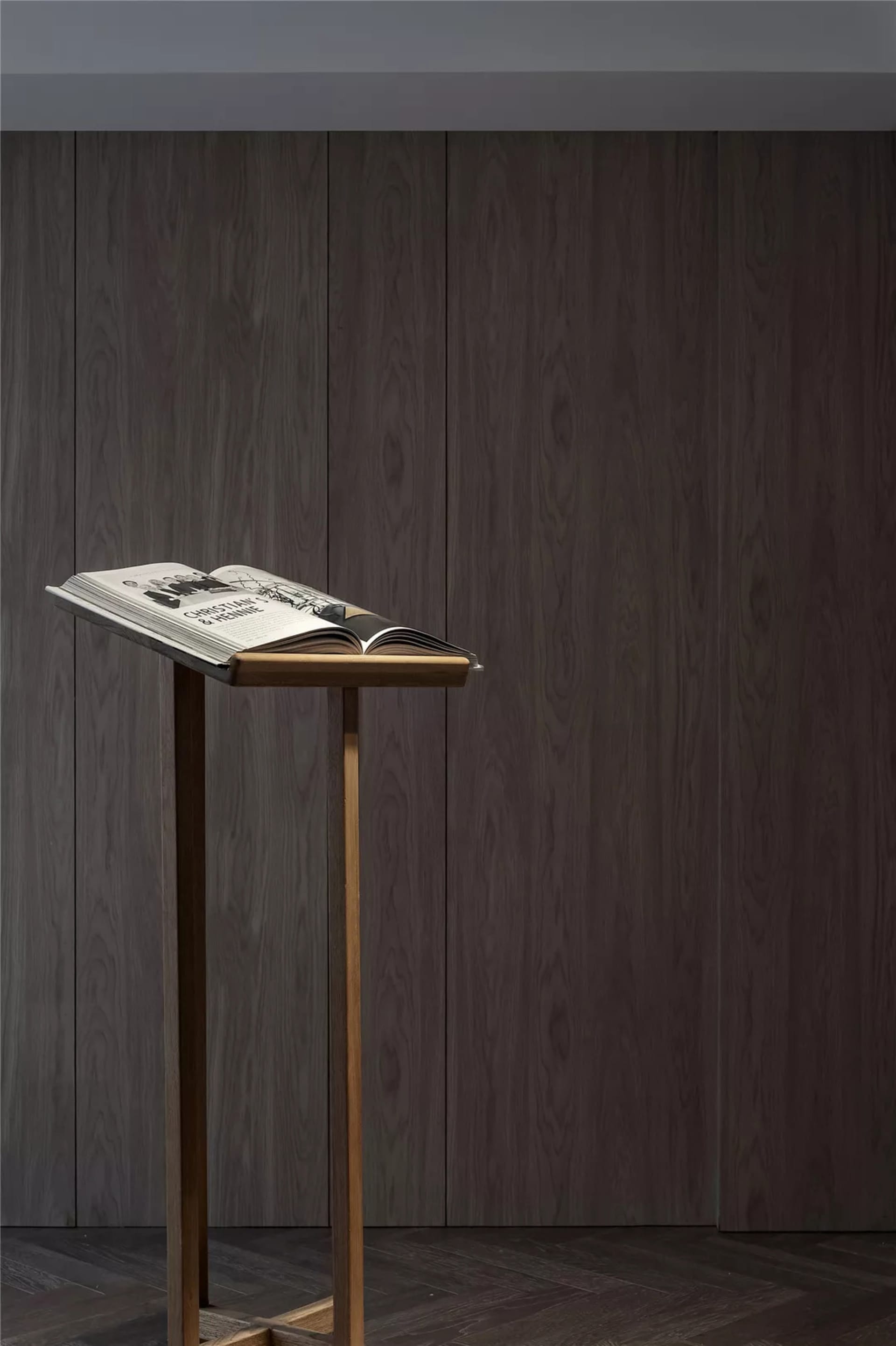 A modern, wooden bookstand holding an open book against a dark, wood-paneled wall.