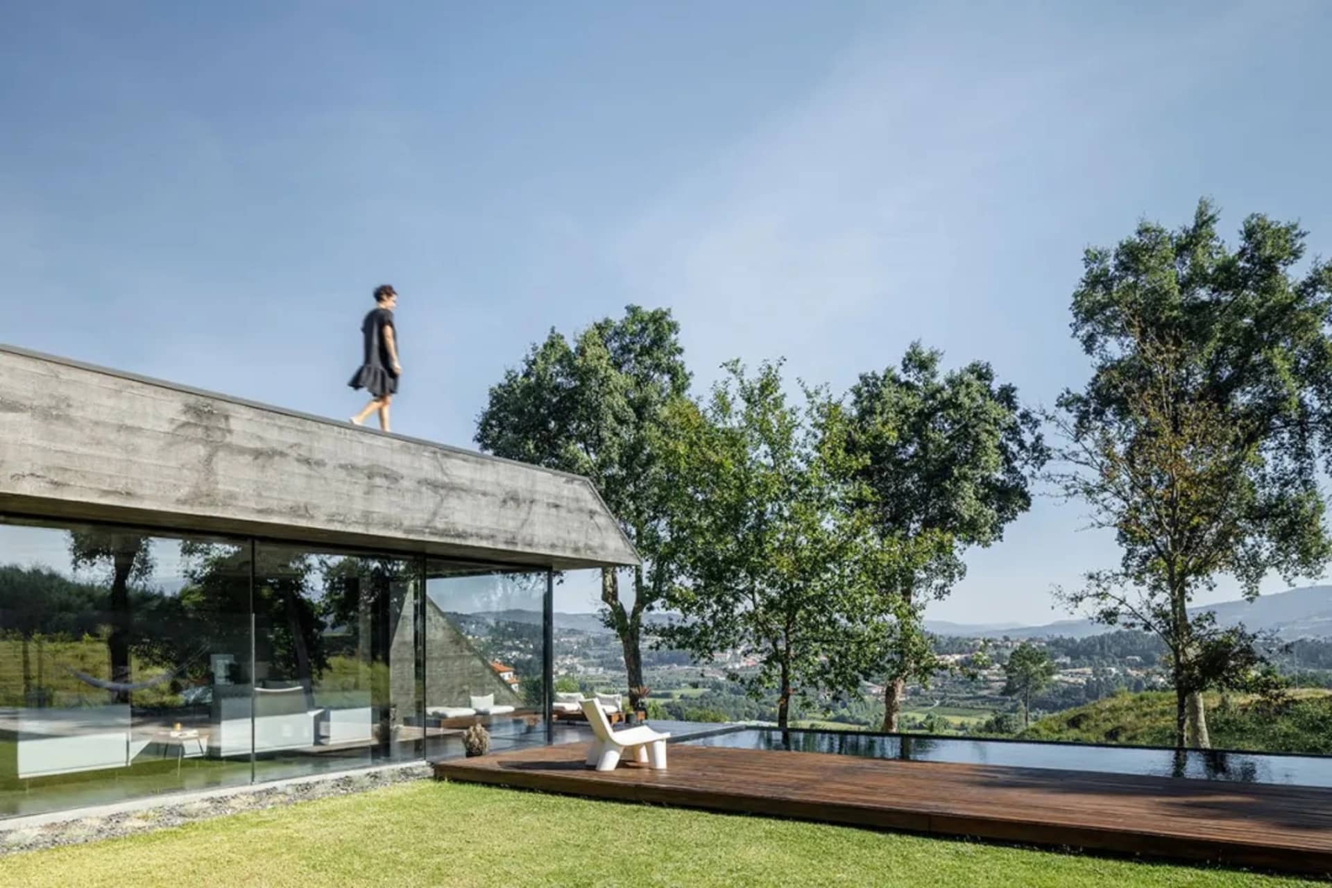A modern house with a concrete roof and glass walls, featuring a person walking on the roof against a clear blue sky.