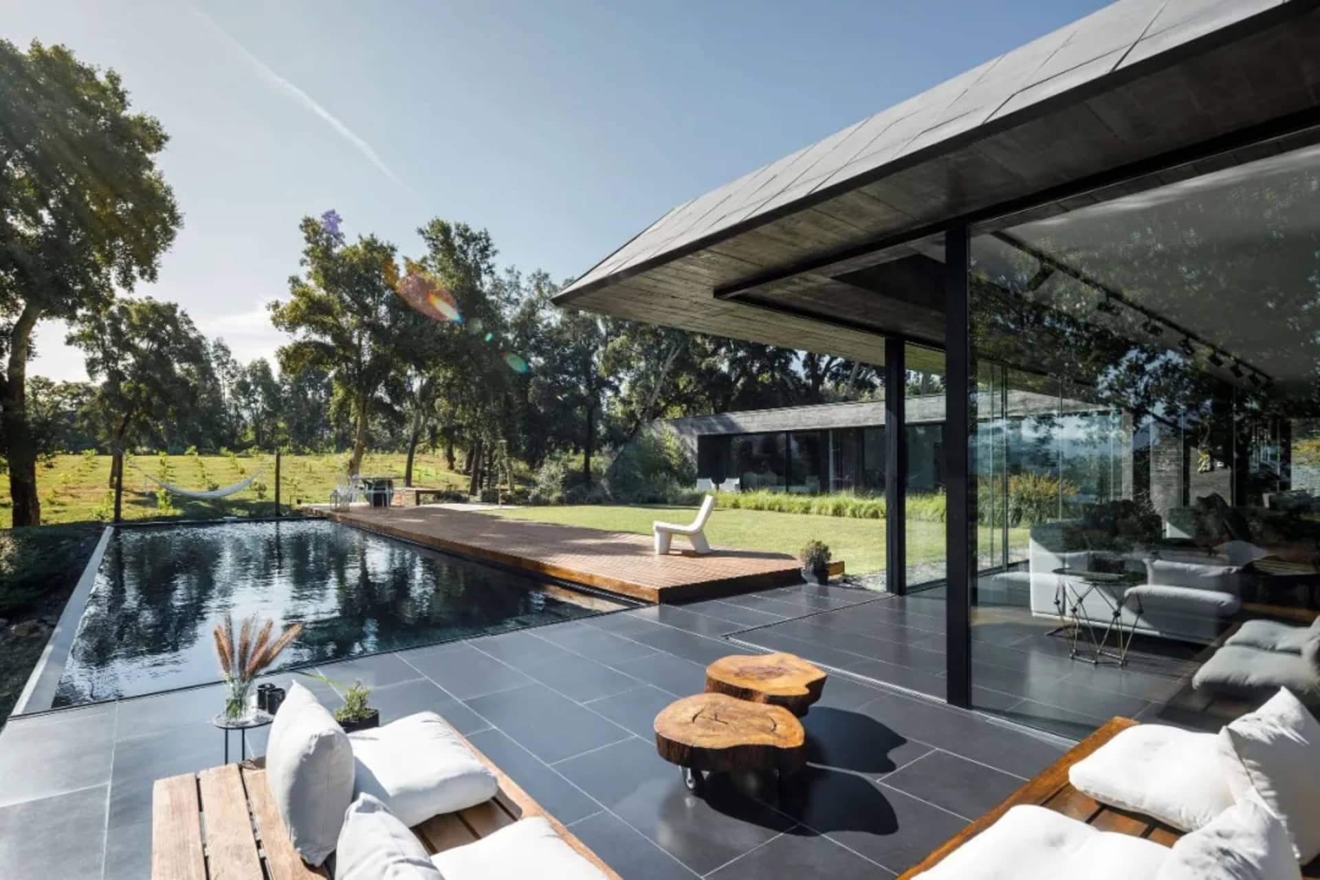 A modern house with a flat roof, glass walls, and concrete patio featuring a swimming pool and outdoor seating area.