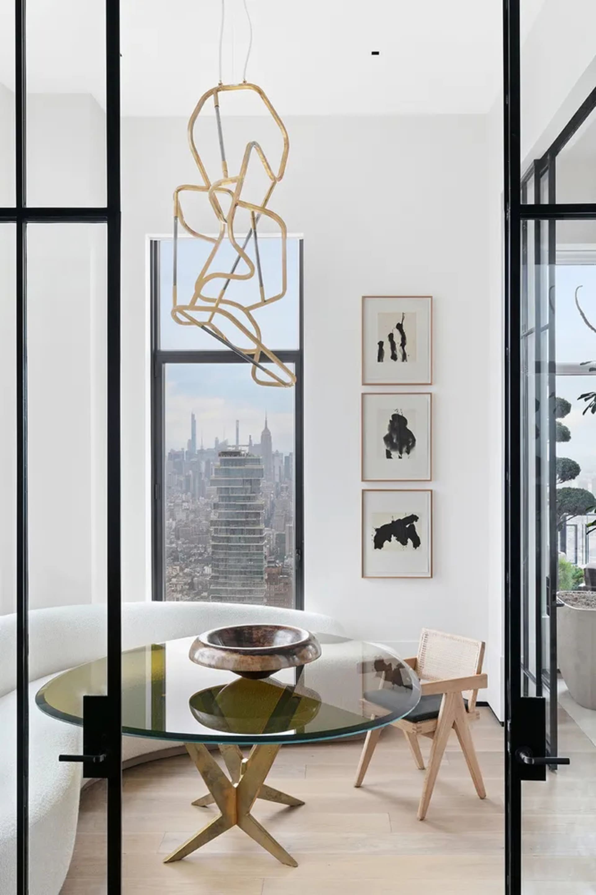 A modern minimalist living room with a glass table, metal chandelier, wooden chair, and steel accents, featuring a cityscape