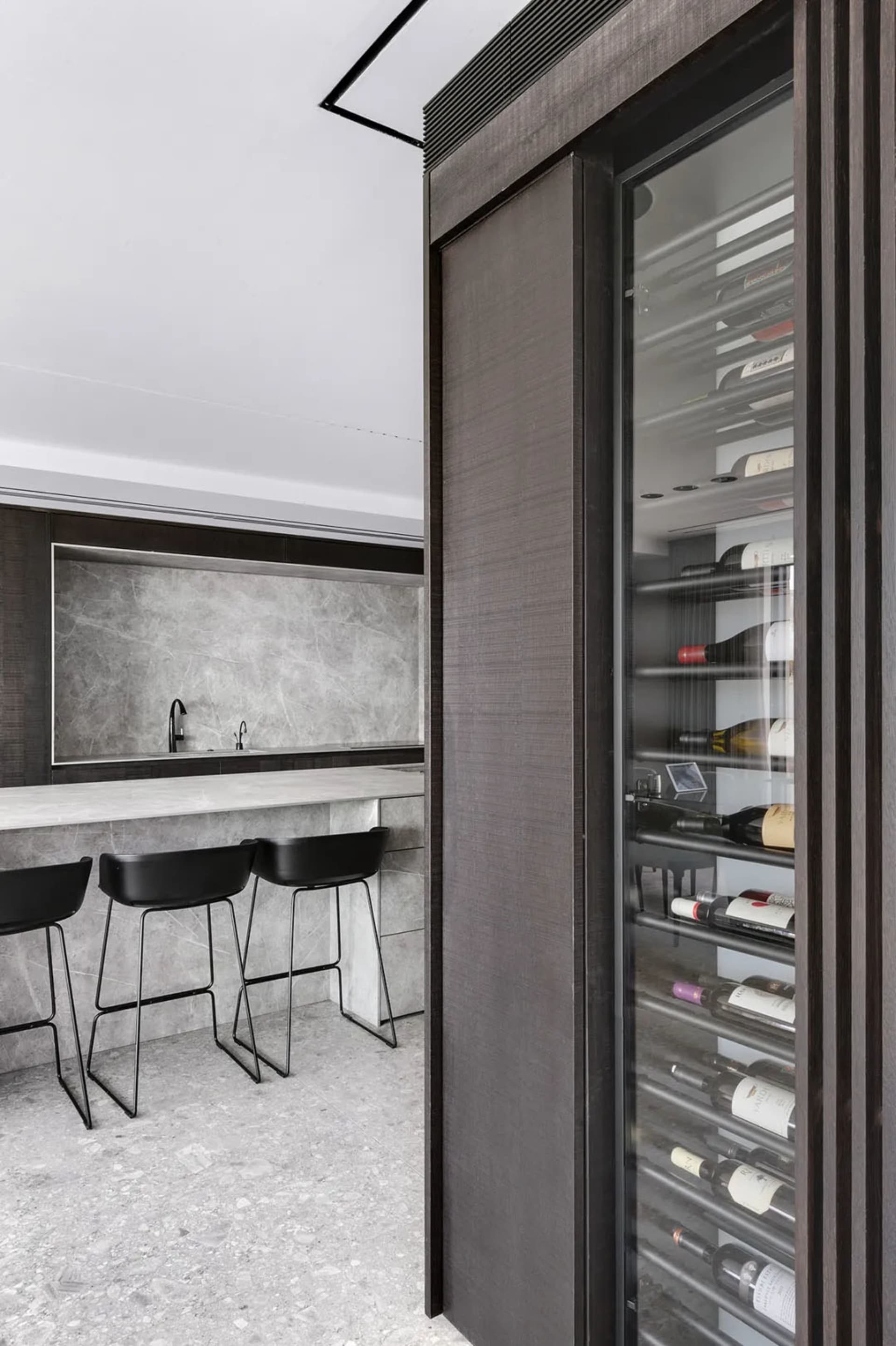 Modern kitchen bar with concrete countertop, black stools, and a glass-fronted wine cabinet.