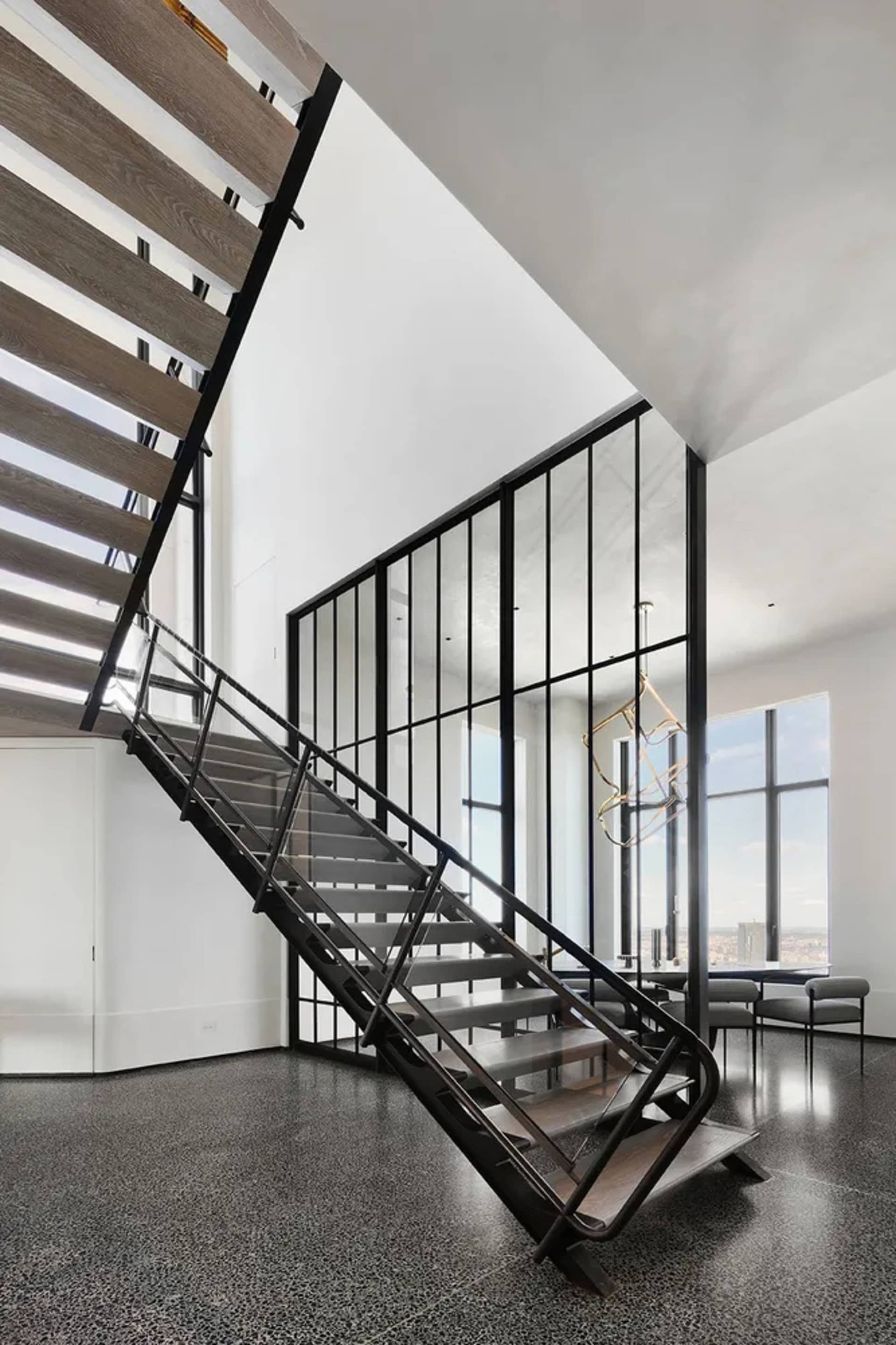 A modern metal staircase with a glass wall and wooden steps, set against a white wall and black floor.