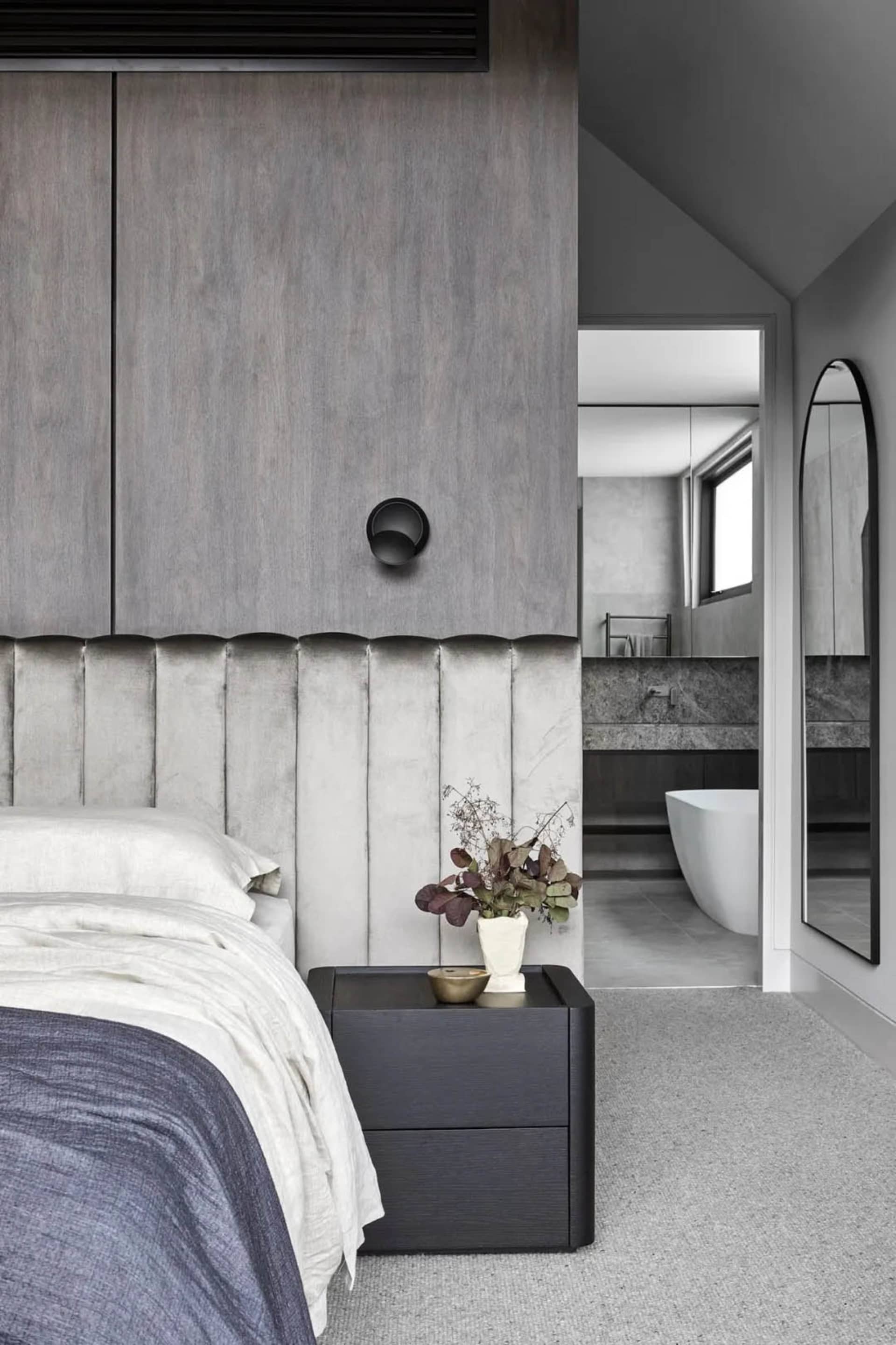 A modern bedroom featuring a gray wall with a textured pattern, a bed with white bedding, and a dark nightstand with a plant.