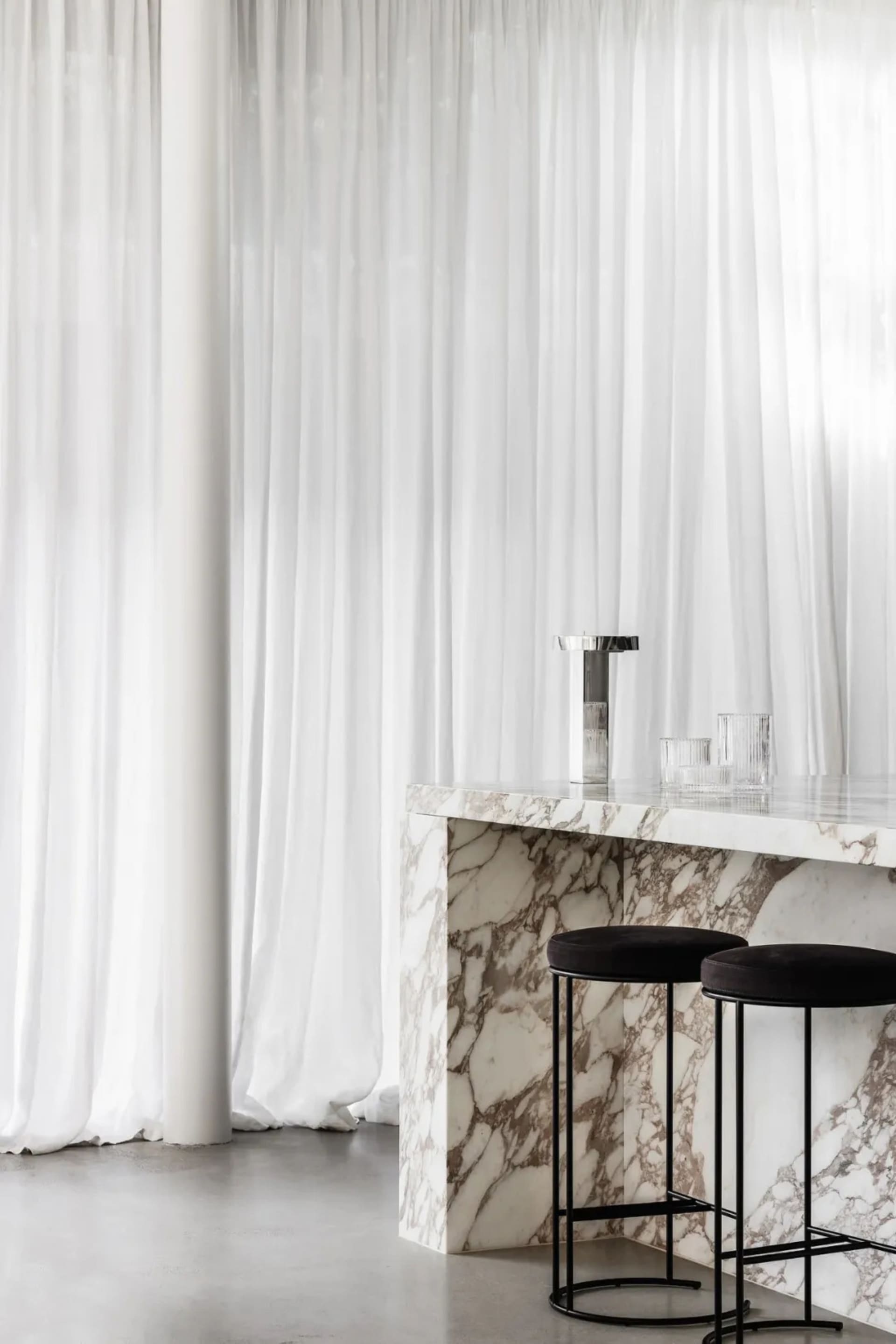 Bar stools placed at a marble counter in front of white curtains