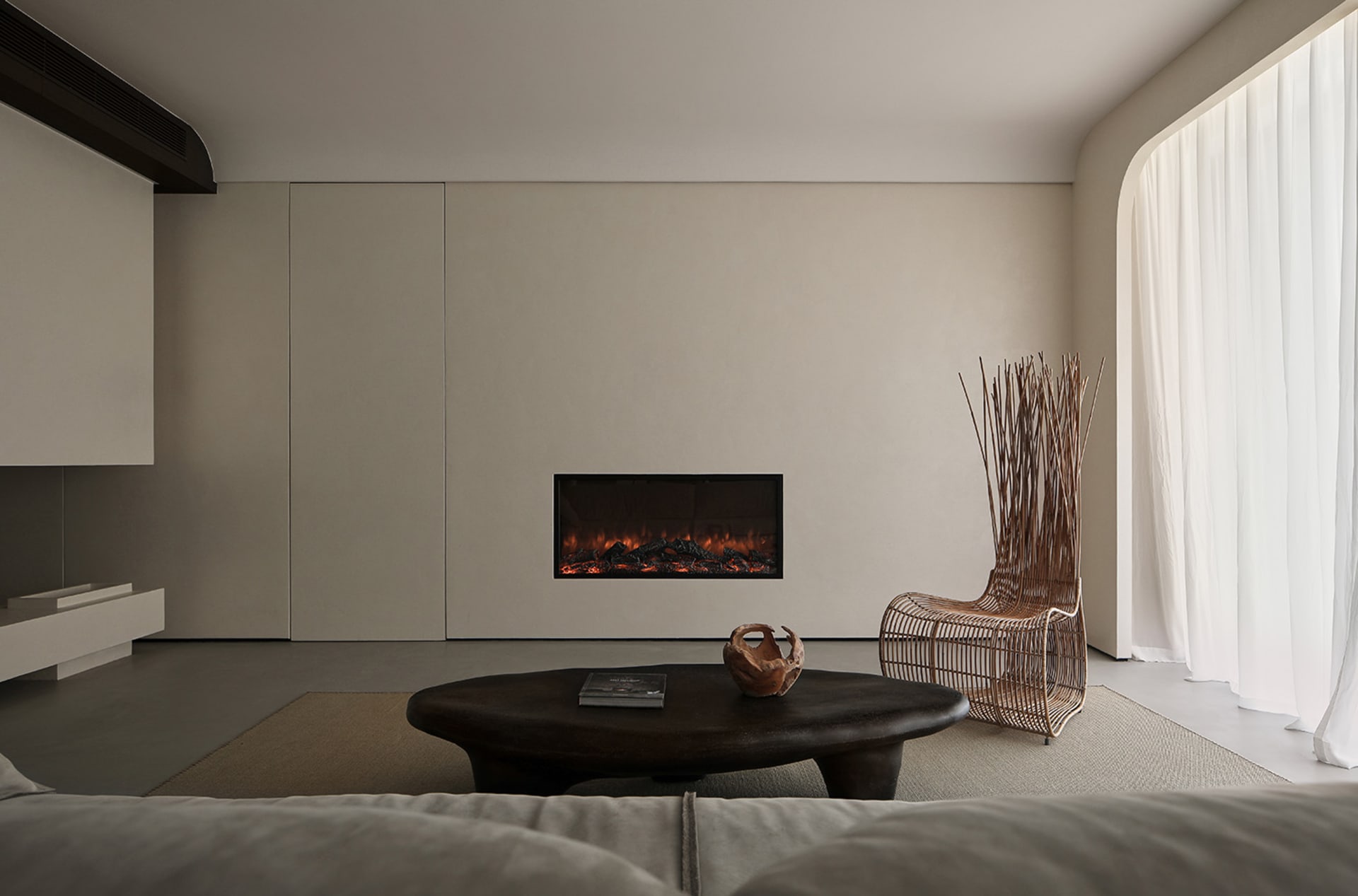 Modern minimalist living room featuring a concrete fireplace, round wooden table, and wire chair, with large windows and soft