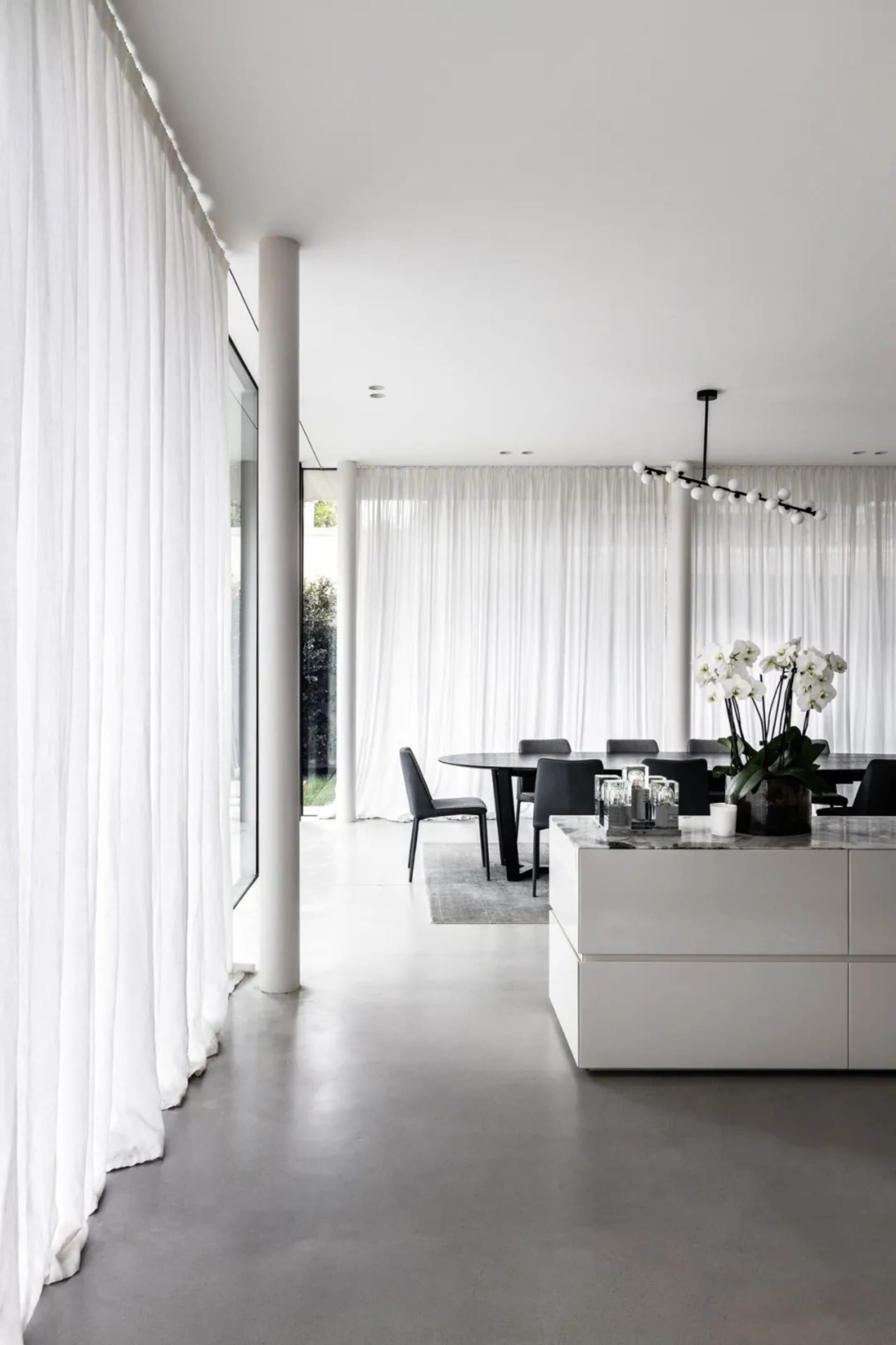 A modern minimalist dining room with white curtains, black furniture, concrete floors, a glass chandelier, and a marble dinin