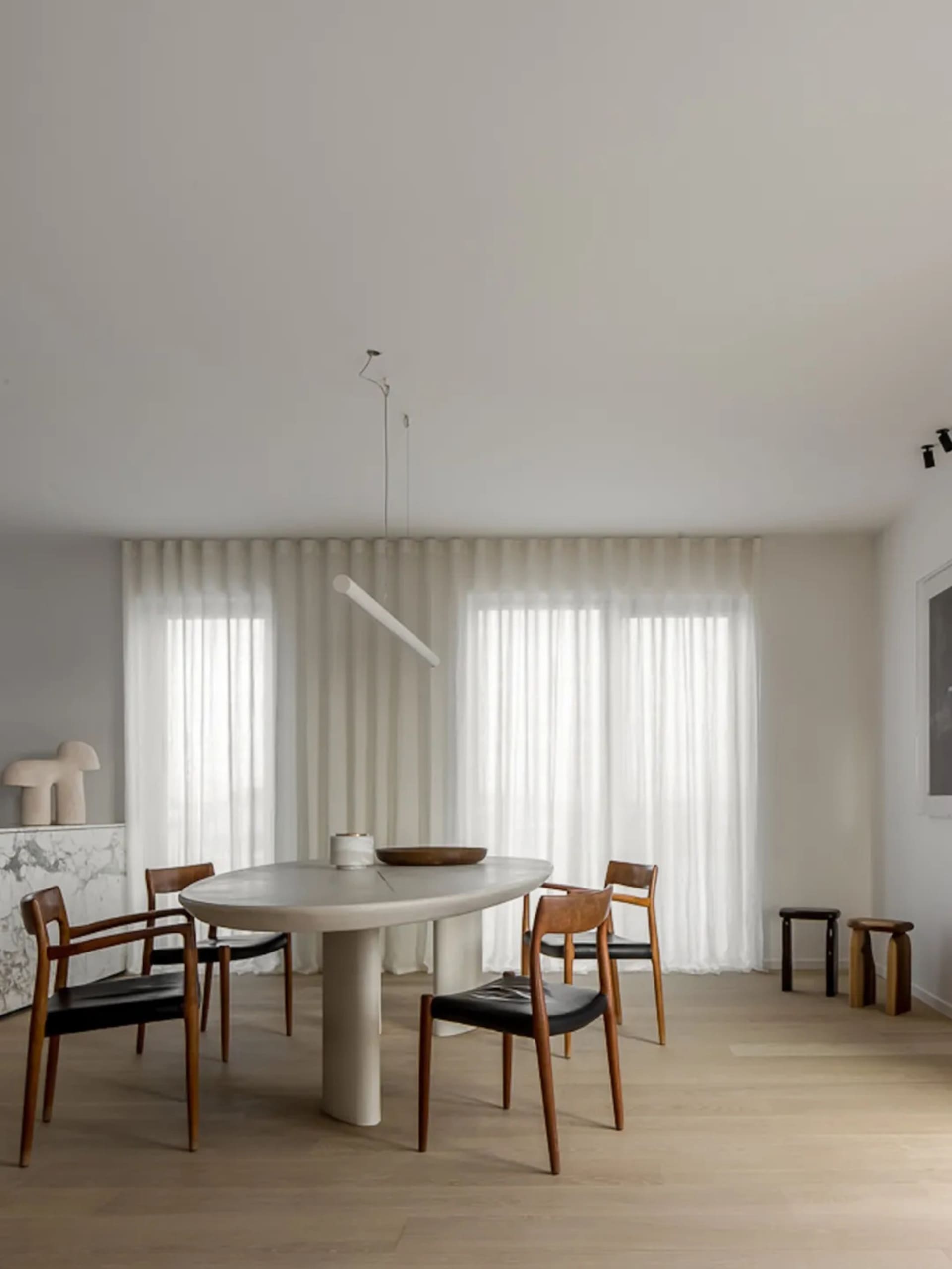 A modern dining room with neutral colors, featuring a minimalist table, wooden chairs, and large windows with sheer curtains.