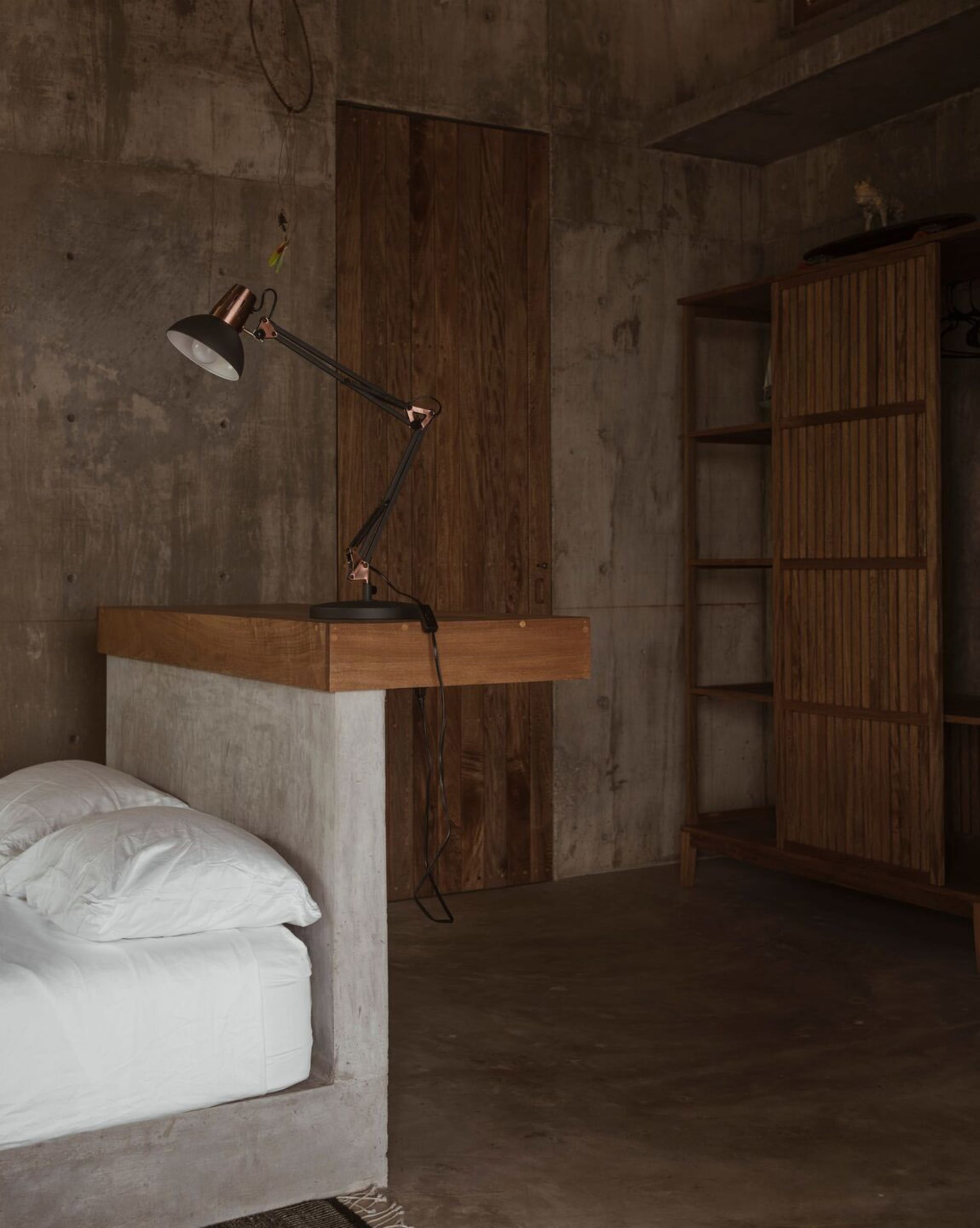 A bedroom with industrial design features a concrete bed frame and wooden desk with a table lamp.