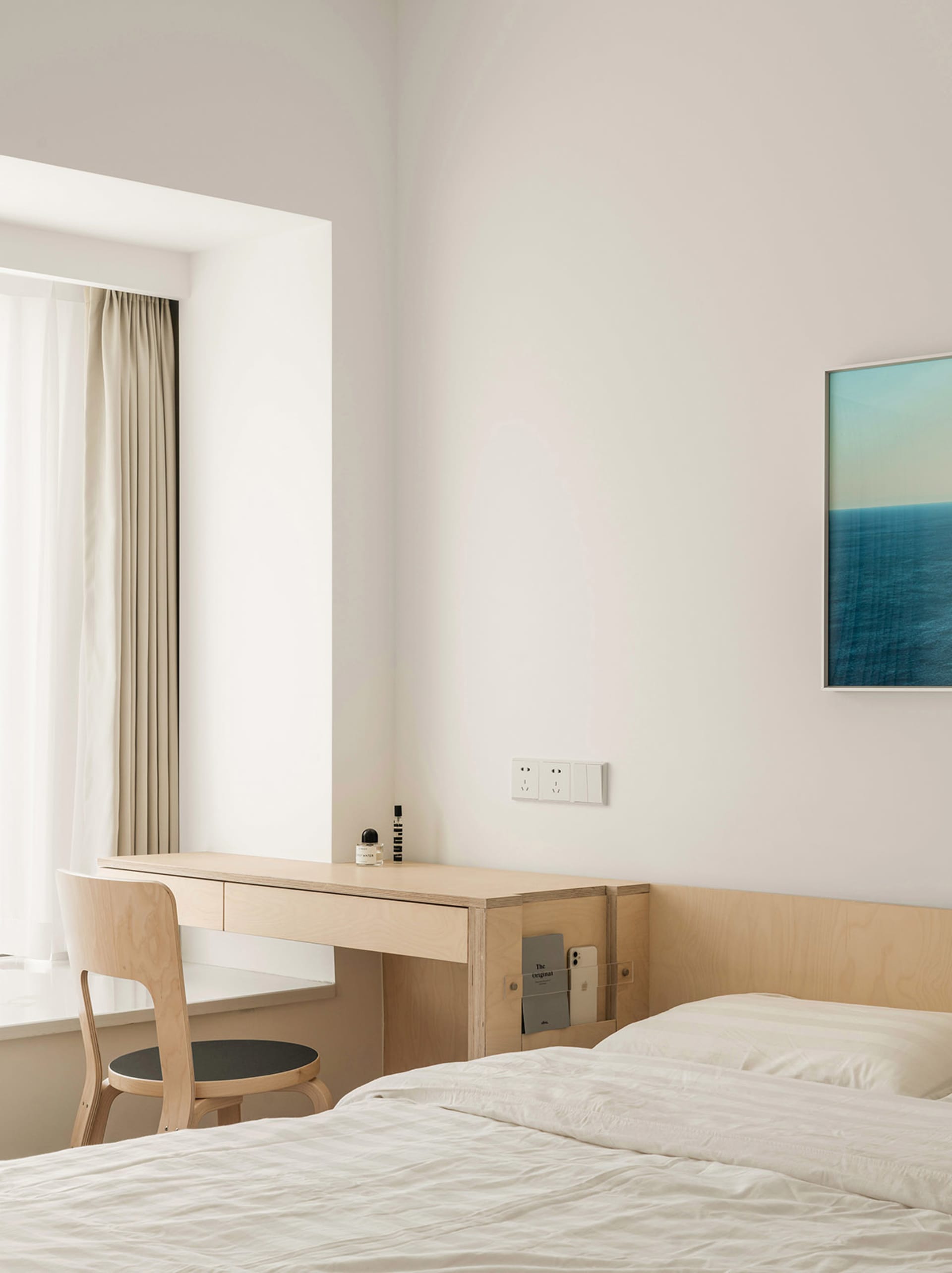 A minimalist bedroom featuring a wooden desk and chair, a bed with white bedding, and a window with light-colored curtains.