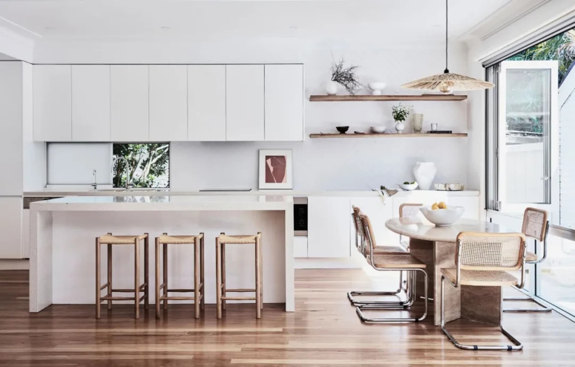 A modern kitchen featuring sleek, white cabinets, wooden stools, and an open shelving unit.