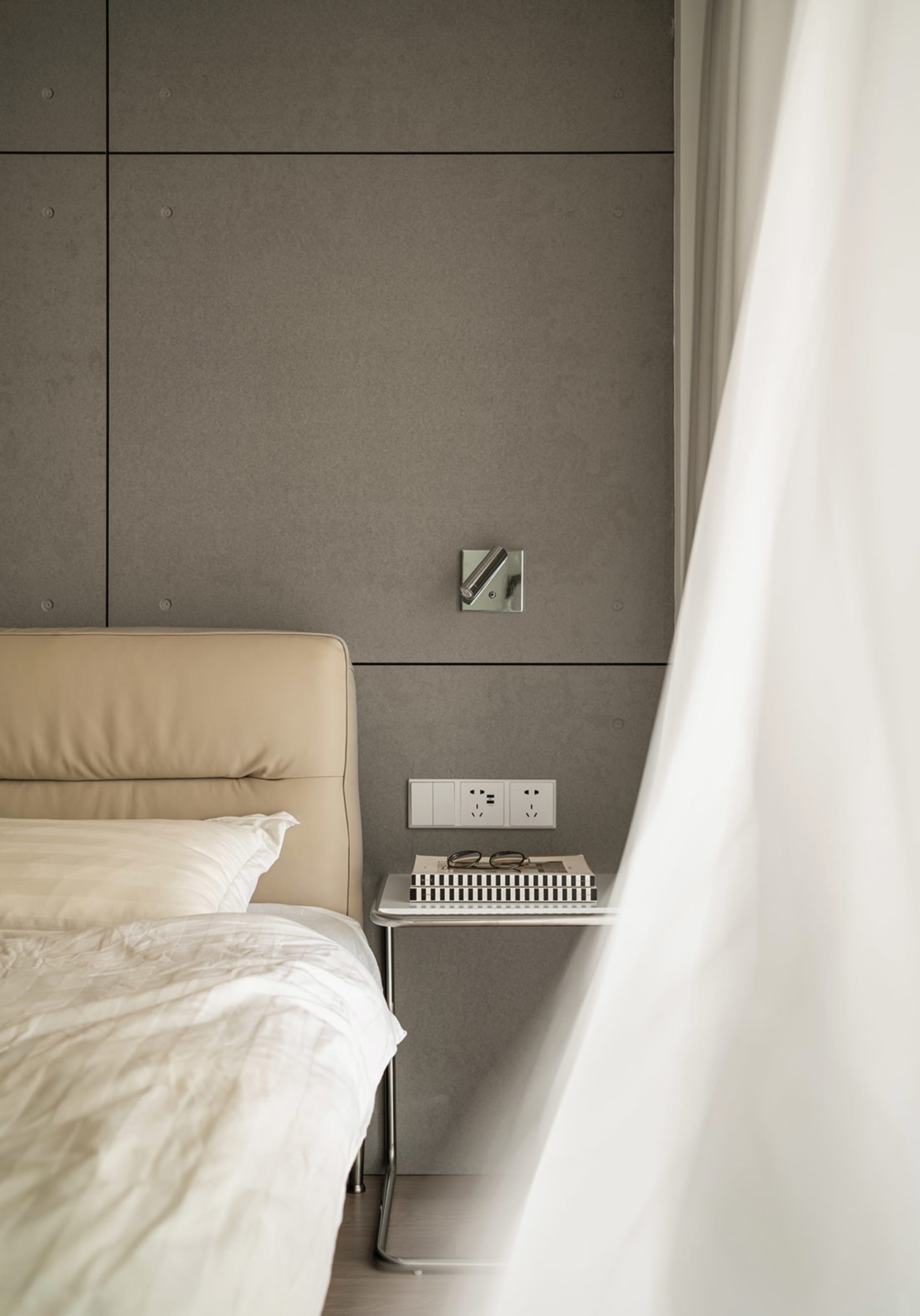 A minimalist bedroom featuring a bed with a beige leather headboard and a nightstand with a book, glasses, and power outlets