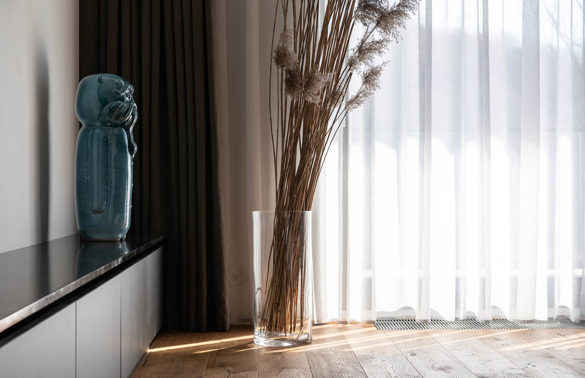 A tall cylindrical glass vase sitting on the floor between curtains and a window, containing dried grasses.