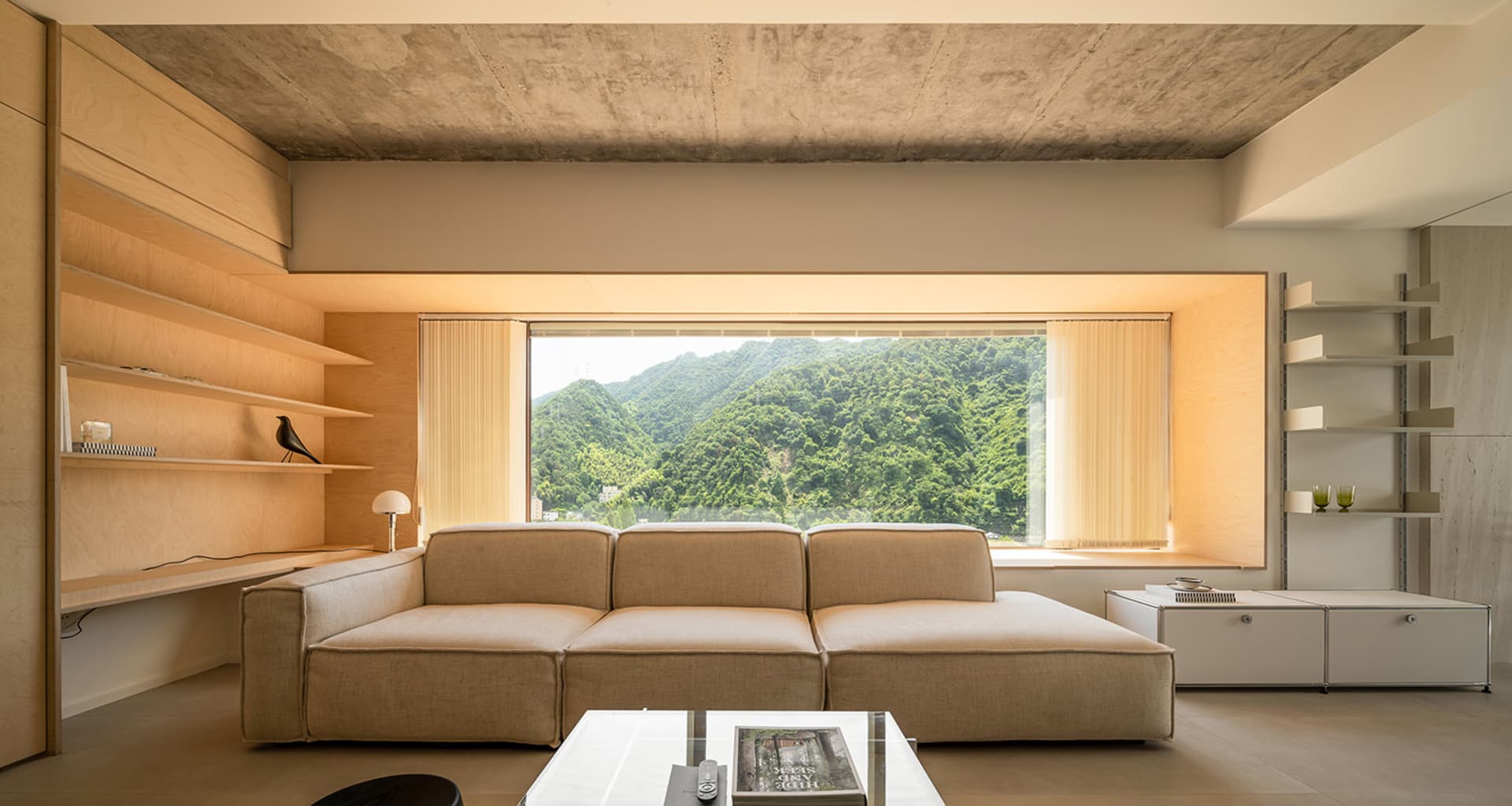 Modern minimalist living room featuring a beige sectional sofa, wooden shelves, concrete ceiling, and a large window overlook