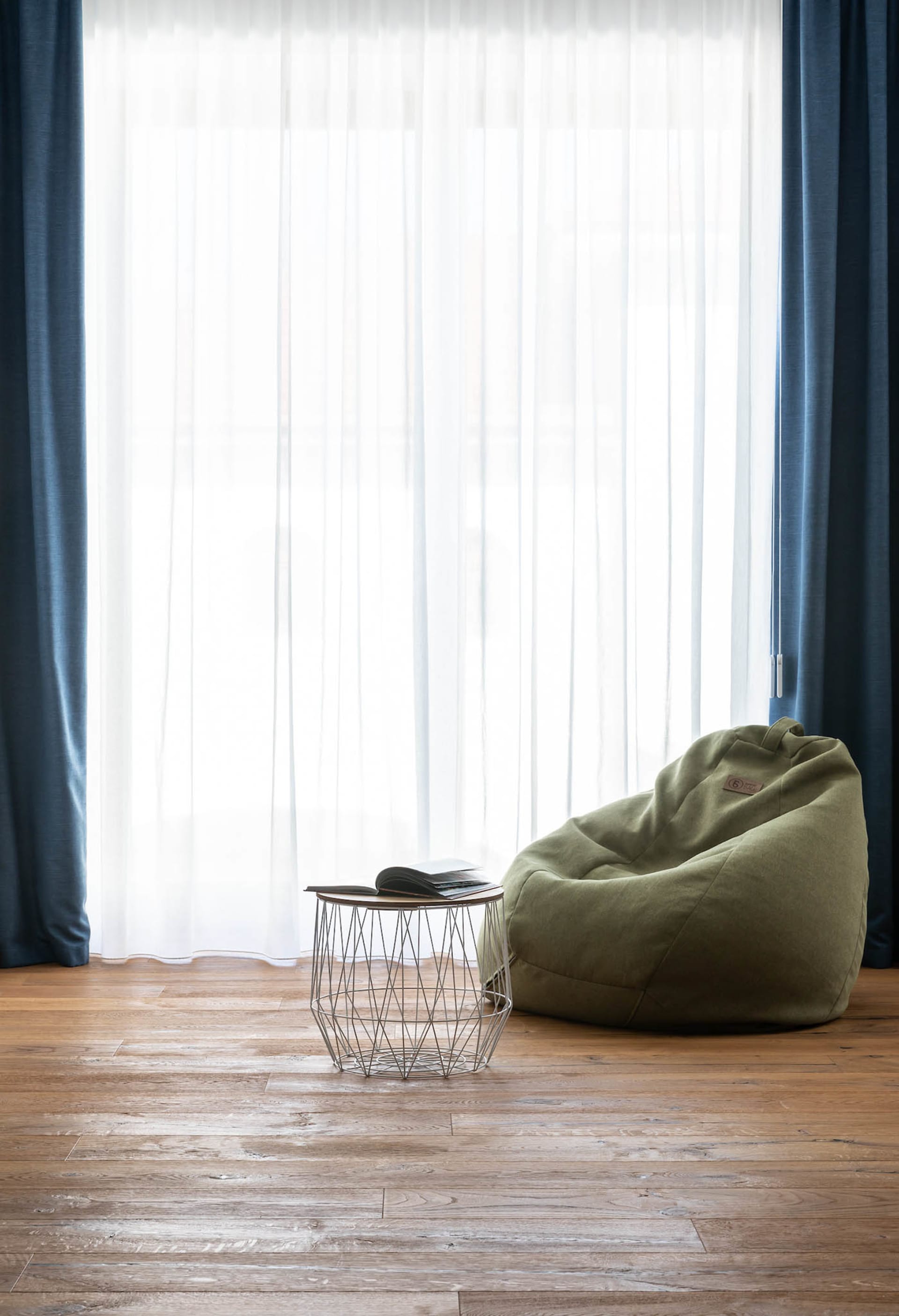 Modern living room featuring a green bean bag chair, a wooden side table with a book, and large windows with sheer curtains,