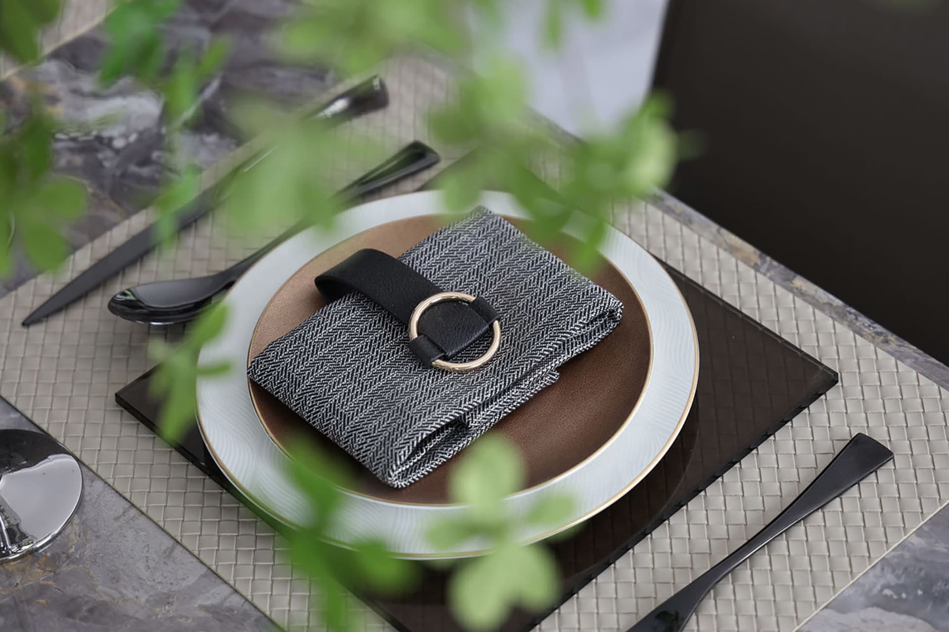 A modern table setting featuring a gray and white place mat, a black leather belt buckle napkin holder, and a gold ring detai
