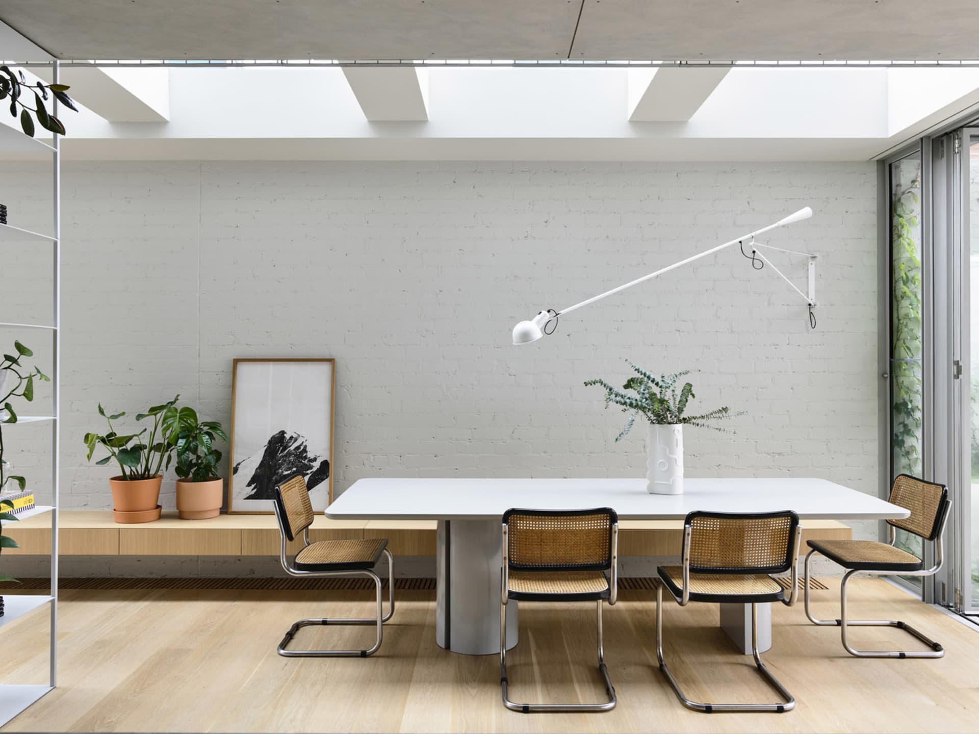 A minimalist dining room with a white table, cane chairs, and a white wall with skylights.