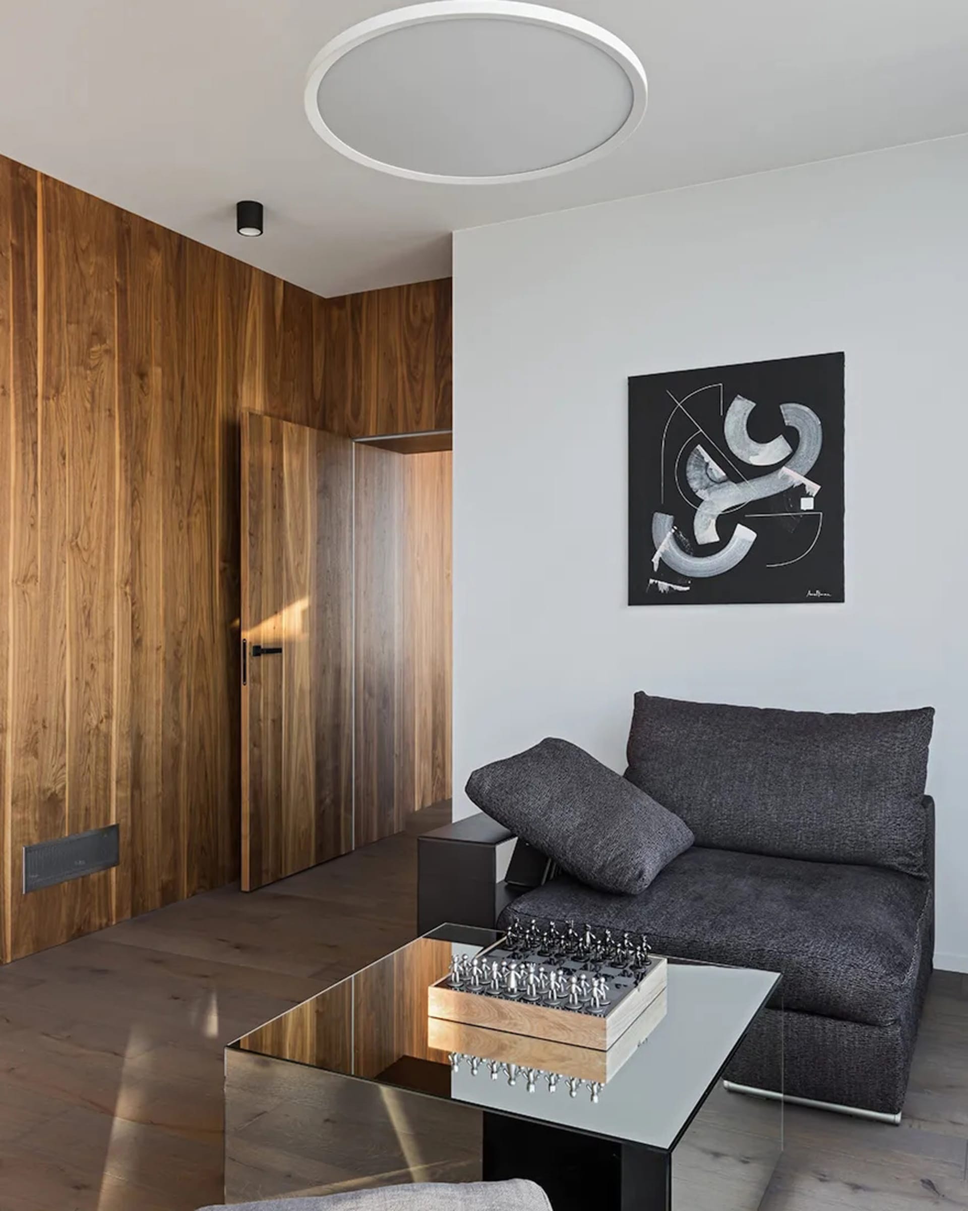 A minimalist living room featuring a wooden wall, glass coffee table, and a grey armless sofa with a mounted artwork.