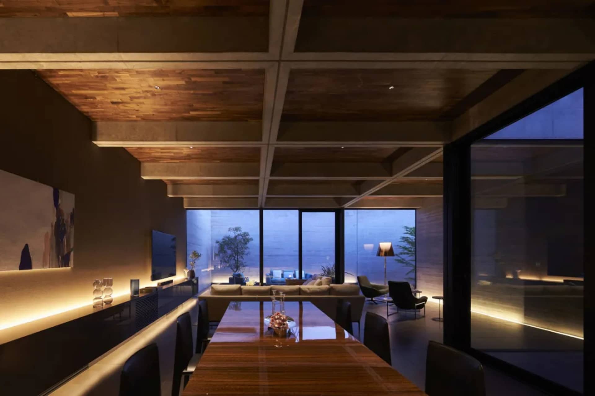 Well-lit dining room with wood and concrete elements, featuring a long table, chairs, and a view of the living area through l