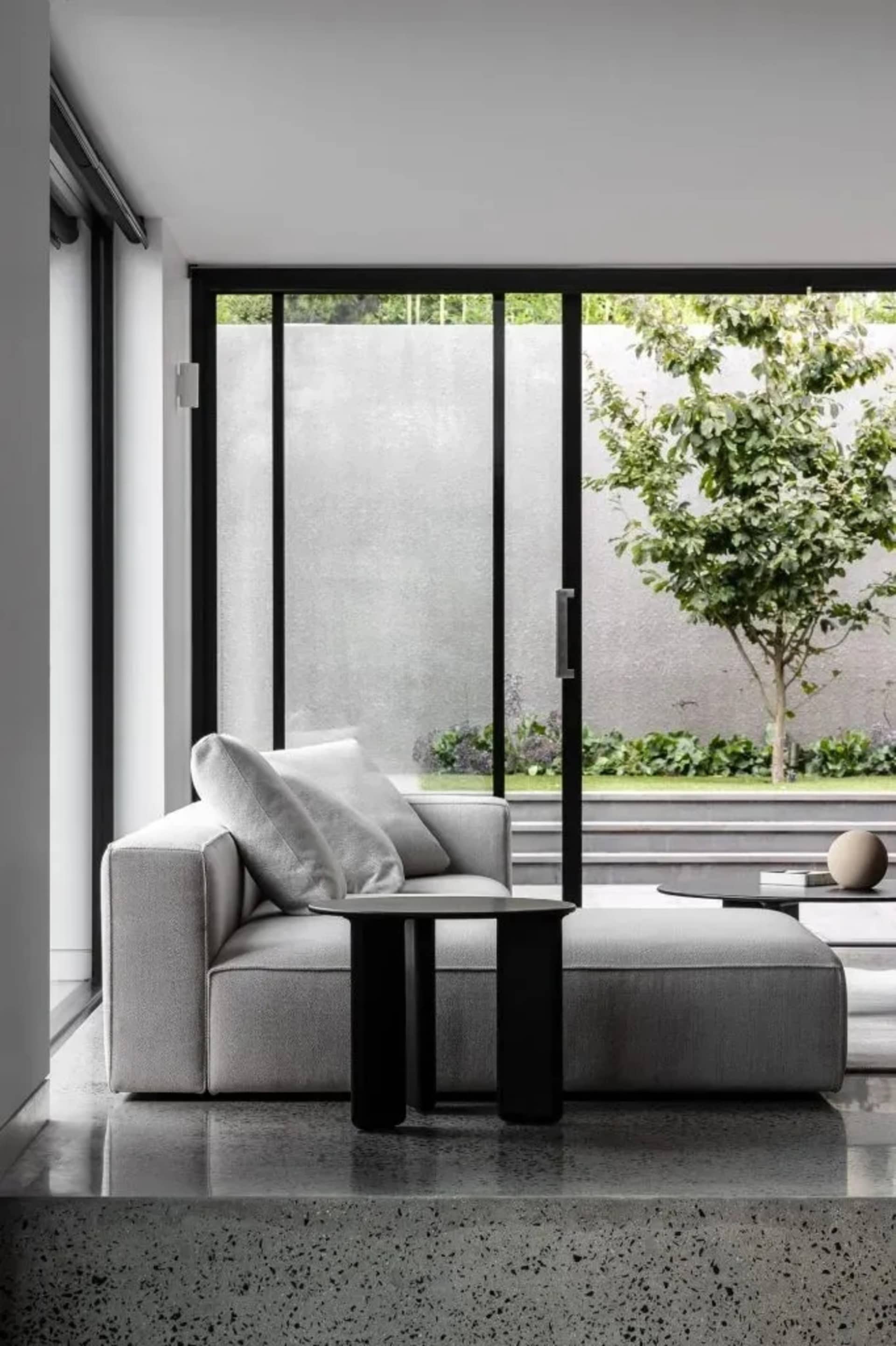 A minimalist living room with a gray modular sofa, black side table, and a view of a tree through large windows.