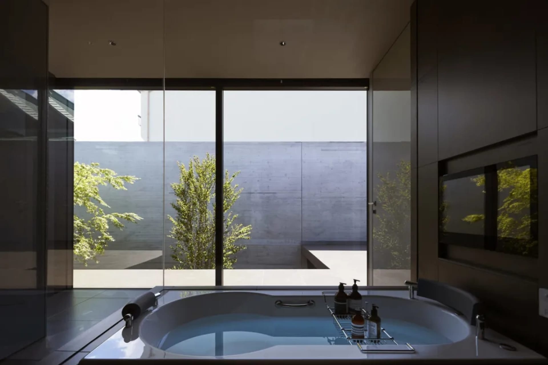 A clean, minimalist bathroom featuring a bathtub with a glass wall, concrete walls, and porcelain tiling.