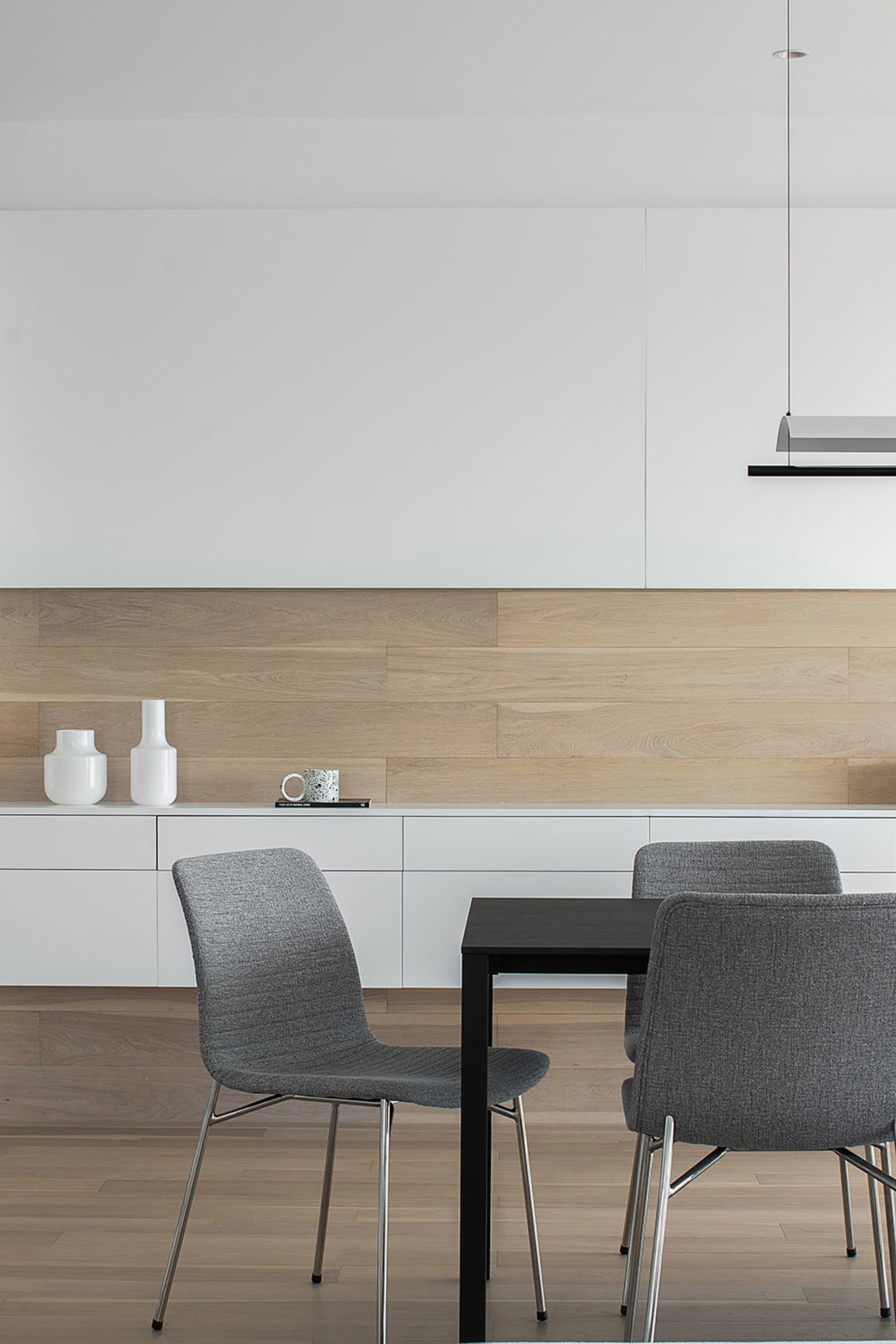 A modern dining room featuring a black table, grey upholstered chairs, and a wooden wall with white floating shelves.