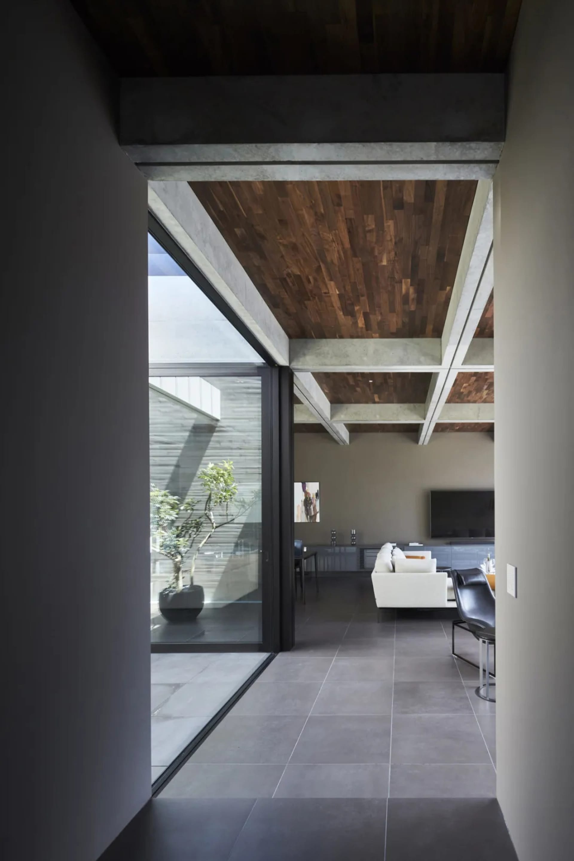 A modern minimalist living room with concrete flooring, wooden ceiling beams, and large glass windows.