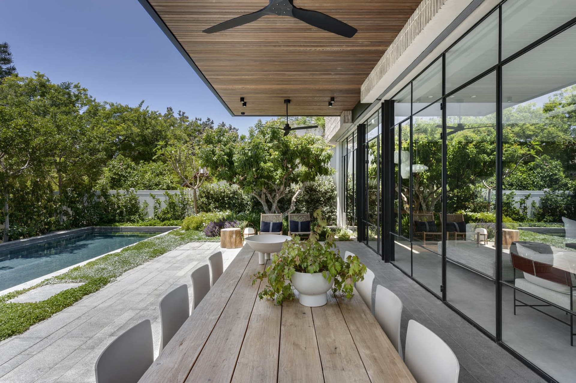 Modern patio area with a wooden dining table, white chairs, and a green plant, surrounded by a pool and lush greenery.