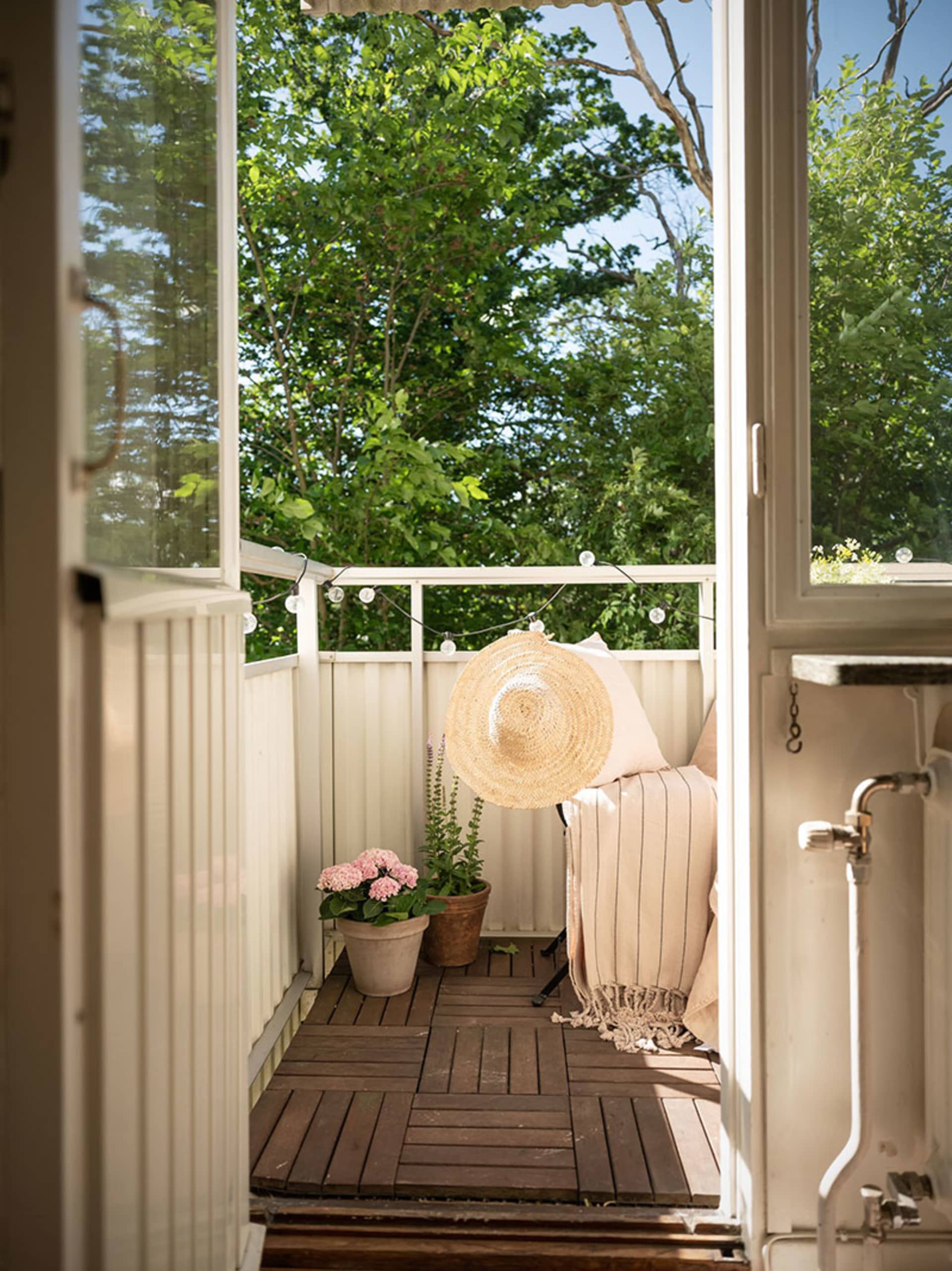 A small balcony features a wooden deck, potted plants, and a cozy sitting area with a beige cushion and throw blanket.