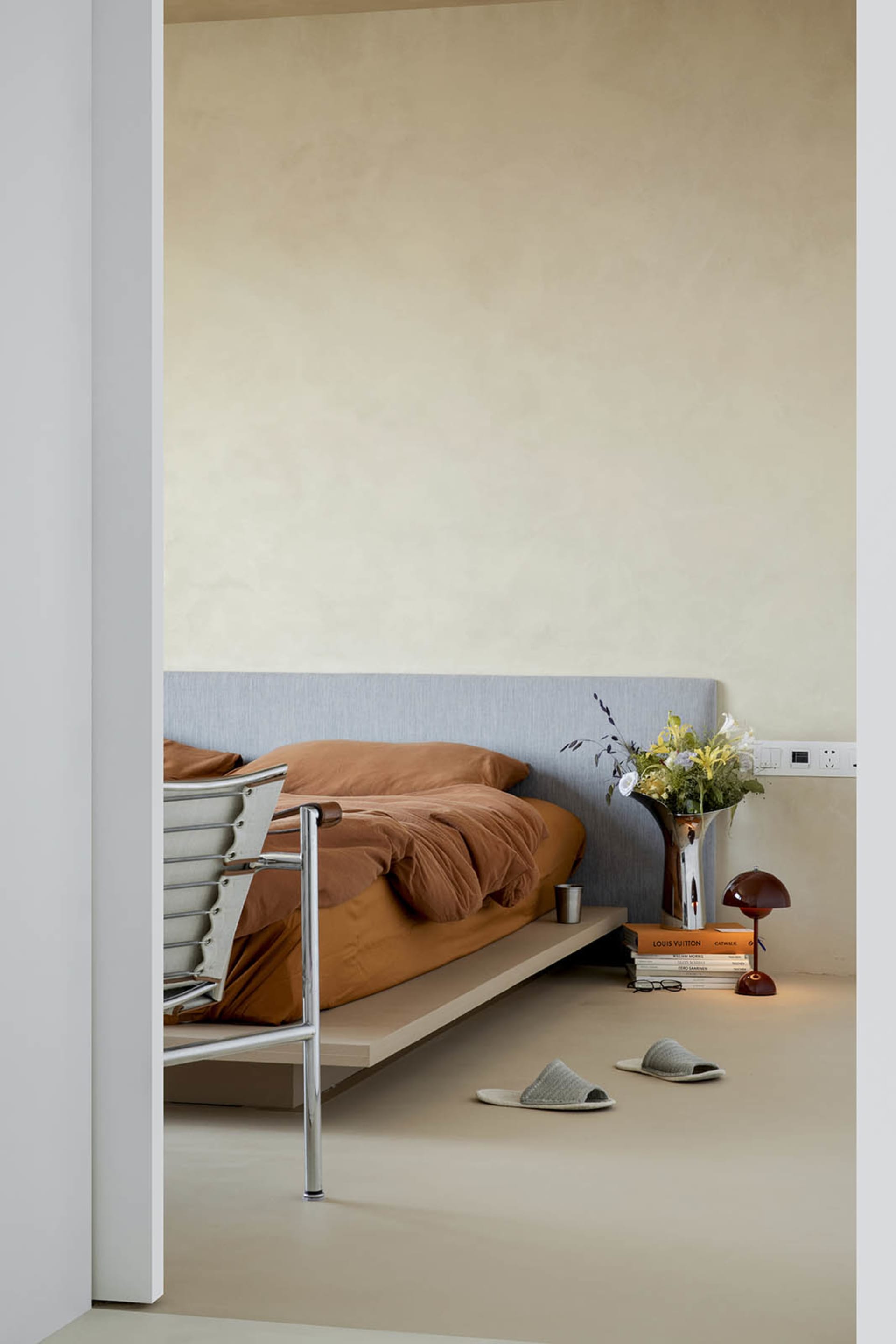 A minimalist bedroom with neutral tones, brown blanket, white slippers, and flowers on the floor