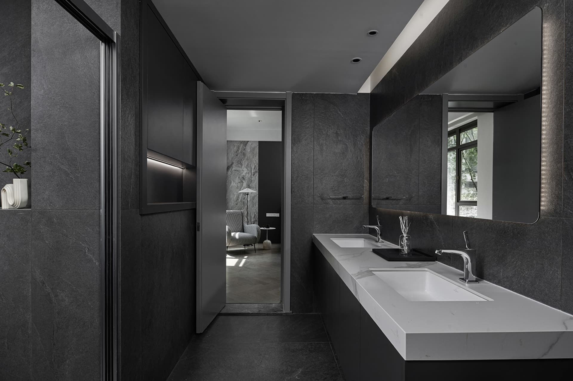 A modern bathroom with black and gray color scheme, white marble sinks, and dark stone walls.