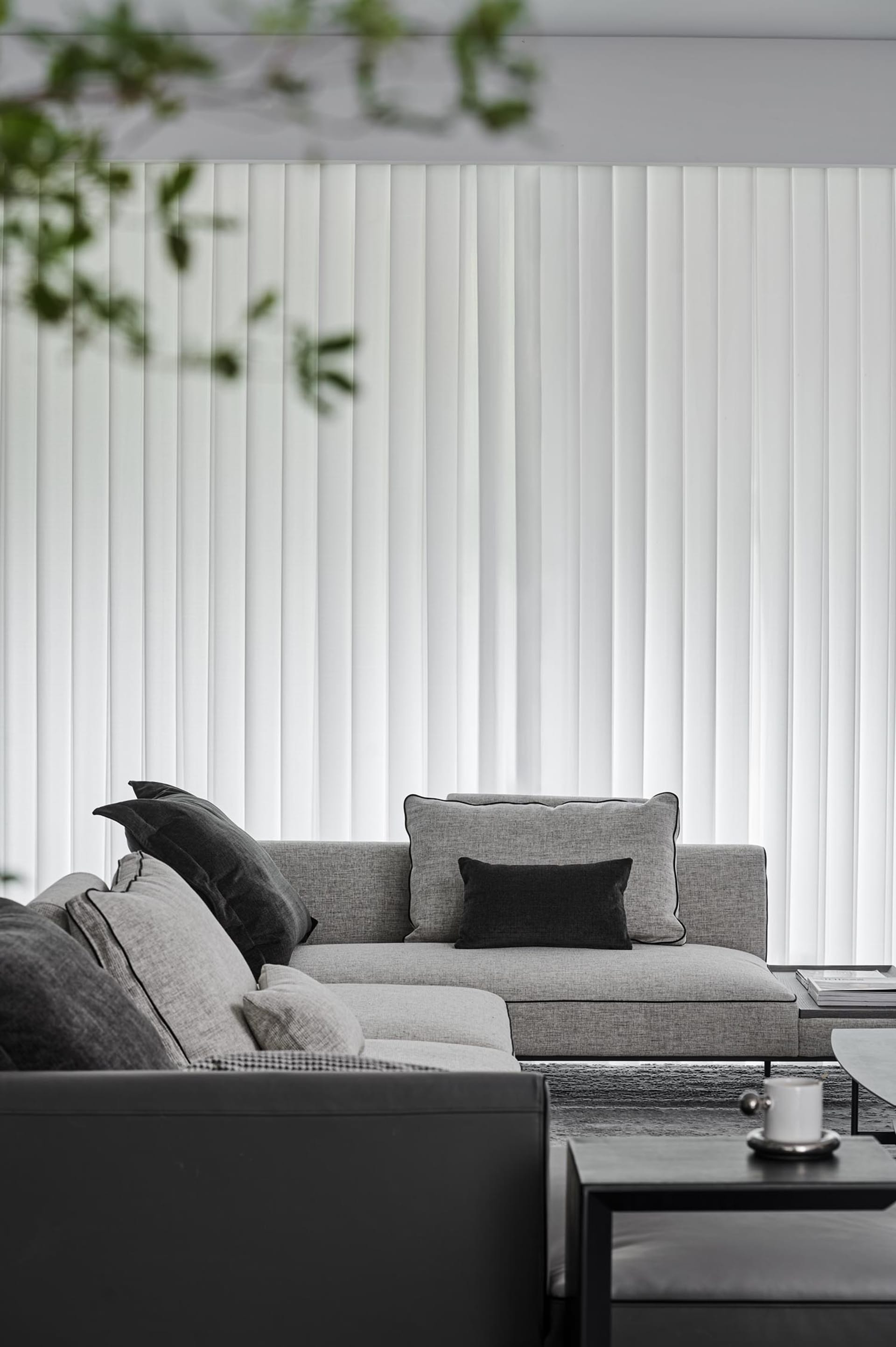 A modern living room featuring a light gray sofa with black accent cushions and white vertical blinds.