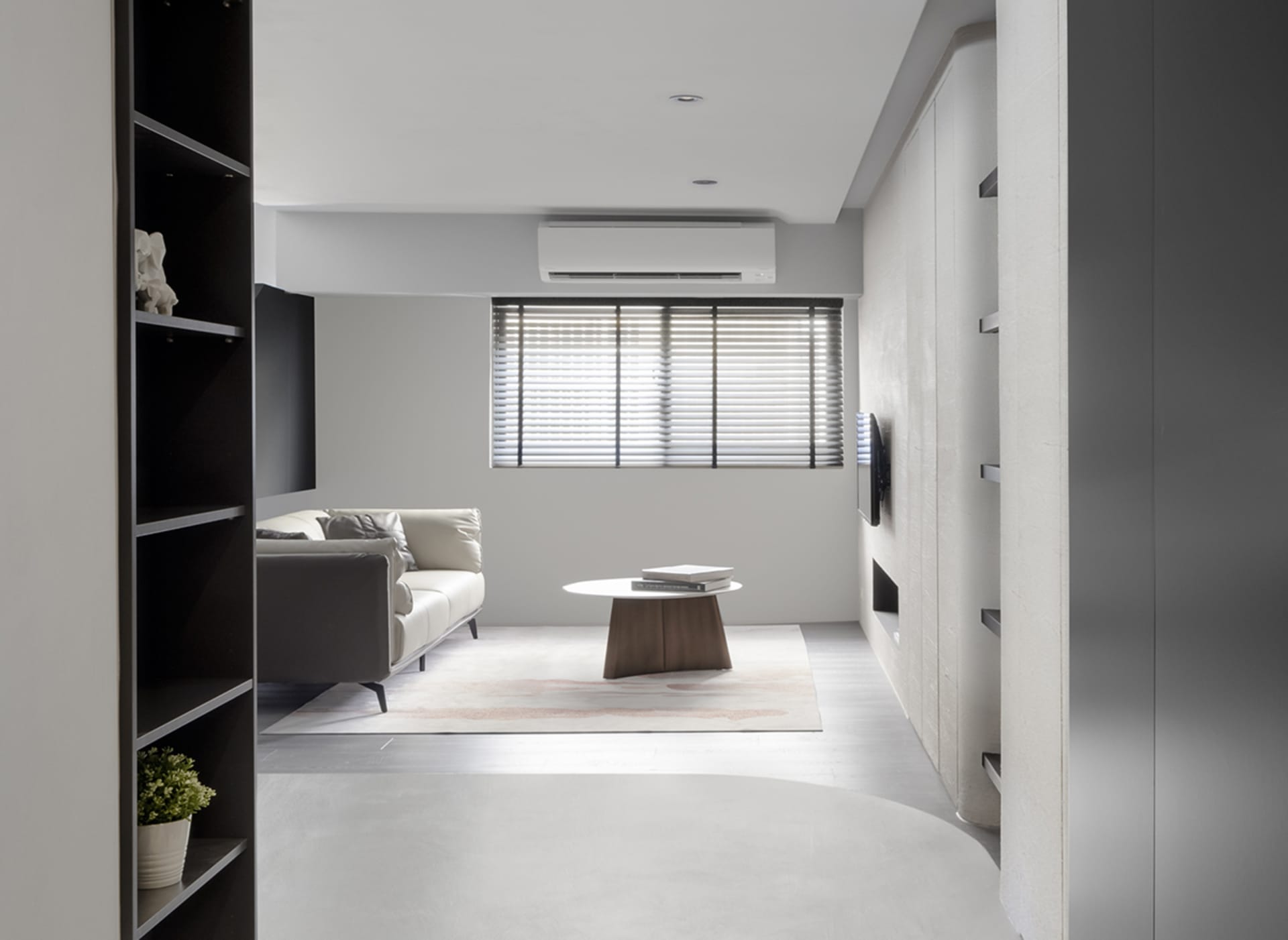 A modern minimalist living room with concrete flooring, a leather sofa, wooden coffee table, and a bookshelf with a plant.